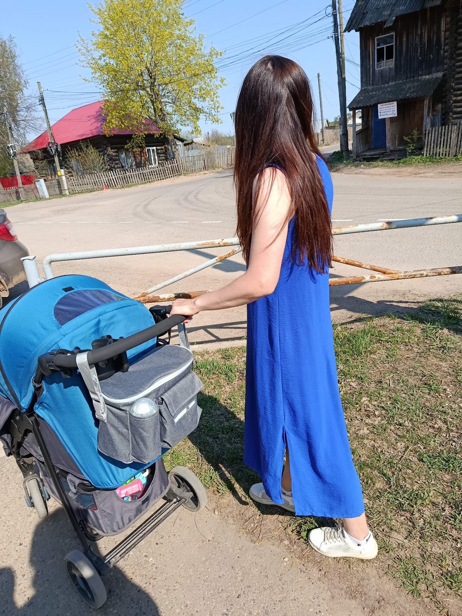 A woman is pushing a blue stroller on a dirt path near a road in Фоки, Russia. The woman has long, dark brown hair and is wearing a long blue dress and white sneakers. The stroller has a grey bag attached to it. In the background, there are wooden houses with red roofs, a barrier, a road, and green trees under a clear blue sky. The scene appears to be a sunny day in a rural or suburban area of the city.