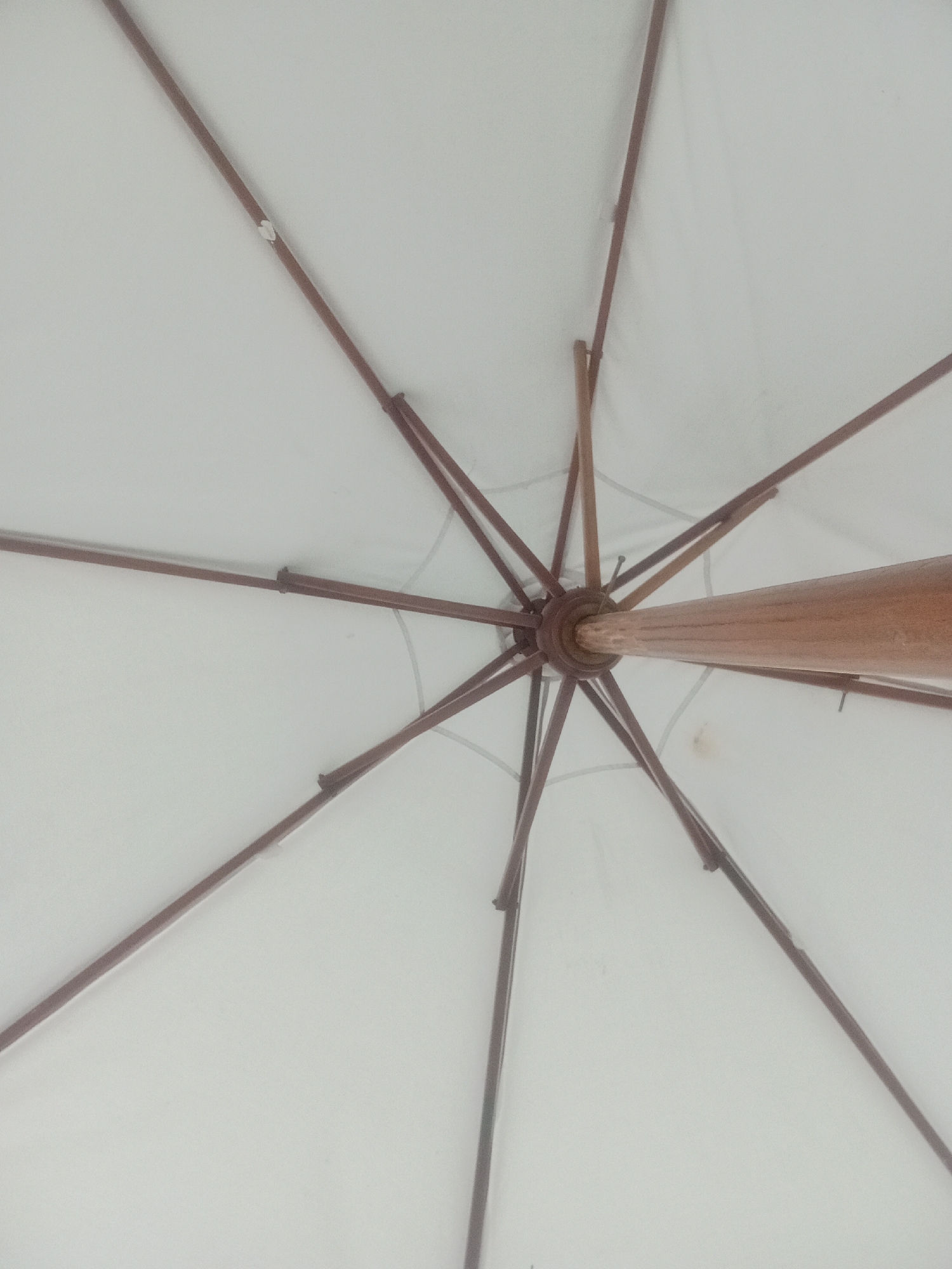 The image is a close-up, overhead view of the underside of a white umbrella. The umbrella has a wooden pole and several thin, brown wooden ribs that radiate outwards from the central pole. The fabric of the umbrella is taut, and the ribs are clearly visible against the white background. There are no people visible in the image. The scene appears to be outdoors, possibly in a resort or beach area, given the context of Kuta Selatan, Indonesia, a region known for its tourist attractions. The overall impression is one of simplicity and functional design.