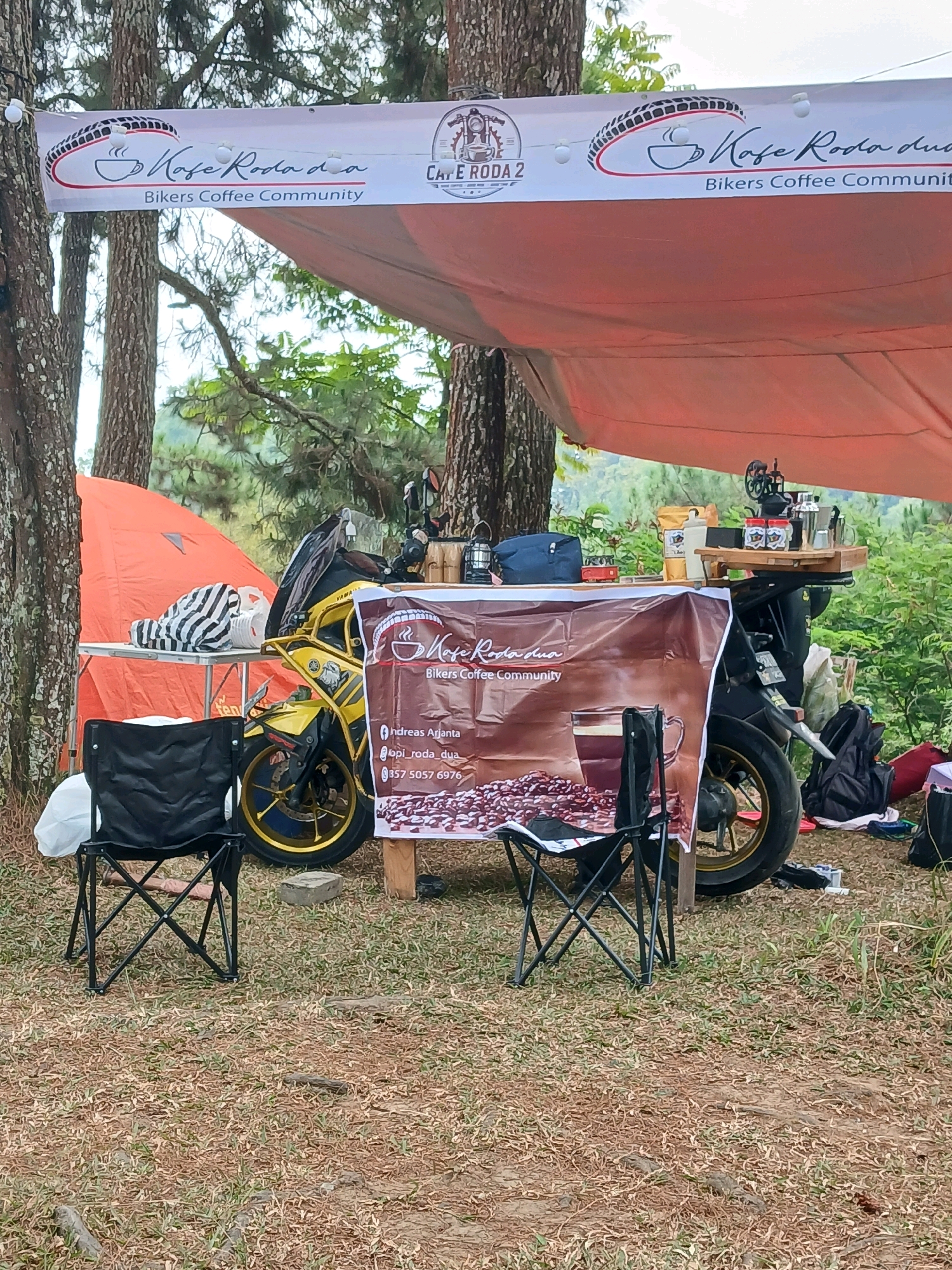 In Berjo, Indonesia, an outdoor coffee stand is set up amidst a wooded area, likely for a biker community event. A yellow motorcycle serves as a central display, with a banner featuring a coffee cup logo and the text "Kafe Roda dua" and "Bikers Coffee Community" stretched between trees. Another banner hangs from a table displaying coffee beans, a coffee cup graphic, and contact information, including an Instagram handle and a phone number. Two black folding chairs are positioned in front of the table, suggesting a seating area for customers. An orange tent and a small table with striped bags are visible to the left, while various coffee brewing equipment and canisters are arranged on a wooden tray atop the motorcycle. The ground is covered in dry grass and dirt.