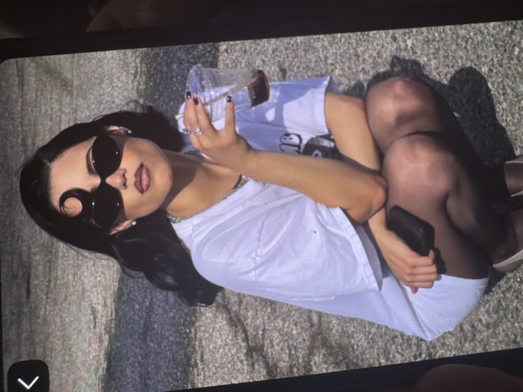 An overhead, slightly angled photograph captures a young woman lying on textured ground, possibly asphalt or concrete, under bright sunlight. The woman is positioned diagonally in the frame, with her head towards the upper left and her legs bent, pointing towards the lower right. She wears large, dark-rimmed sunglasses that obscure her eyes, and her dark hair is spread out beneath her head. Her lips are parted slightly, showing a hint of lipstick.
She is dressed in a loose-fitting white t-shirt, which has an indistinct dark graphic or text visible on the chest area, and what appear to be dark shorts or underwear beneath. Her legs are bent at the knees, revealing bare skin, and she is wearing light-colored shoes, possibly sandals. Her left arm is raised, holding a clear plastic cup containing a dark beverage, likely iced coffee or tea, with a straw. Several rings are visible on the fingers of her left hand, and her fingernails are painted with dark polish. In her right hand, she holds a black smartphone. Strong shadows are cast on the ground from her body, indicating a sunny daytime setting. The overall mood is casual and relaxed. The exact location is not discernible from the image.