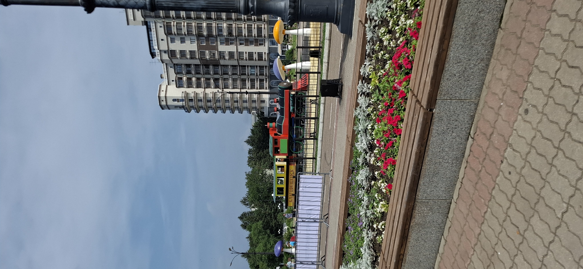 The image depicts a public park area in Blagoveshchensk, Russia, featuring a decorative train and children's playground elements. In the foreground, a well-maintained flower bed with red and white flowers is bordered by wooden benches and a paved walkway. Behind the flower bed, a decorative fence separates the park area from a street. A colorful, stationary train, designed as a children's play structure, is visible, with Cyrillic text on its side. Two large, stylized mushroom figures, one blue and one orange, stand near the train. In the background, a tall, modern apartment building with multiple balconies dominates the skyline. The sky is partly cloudy. Several individuals are present in the background, appearing to be visitors to the park. The overall scene suggests a recreational urban space.