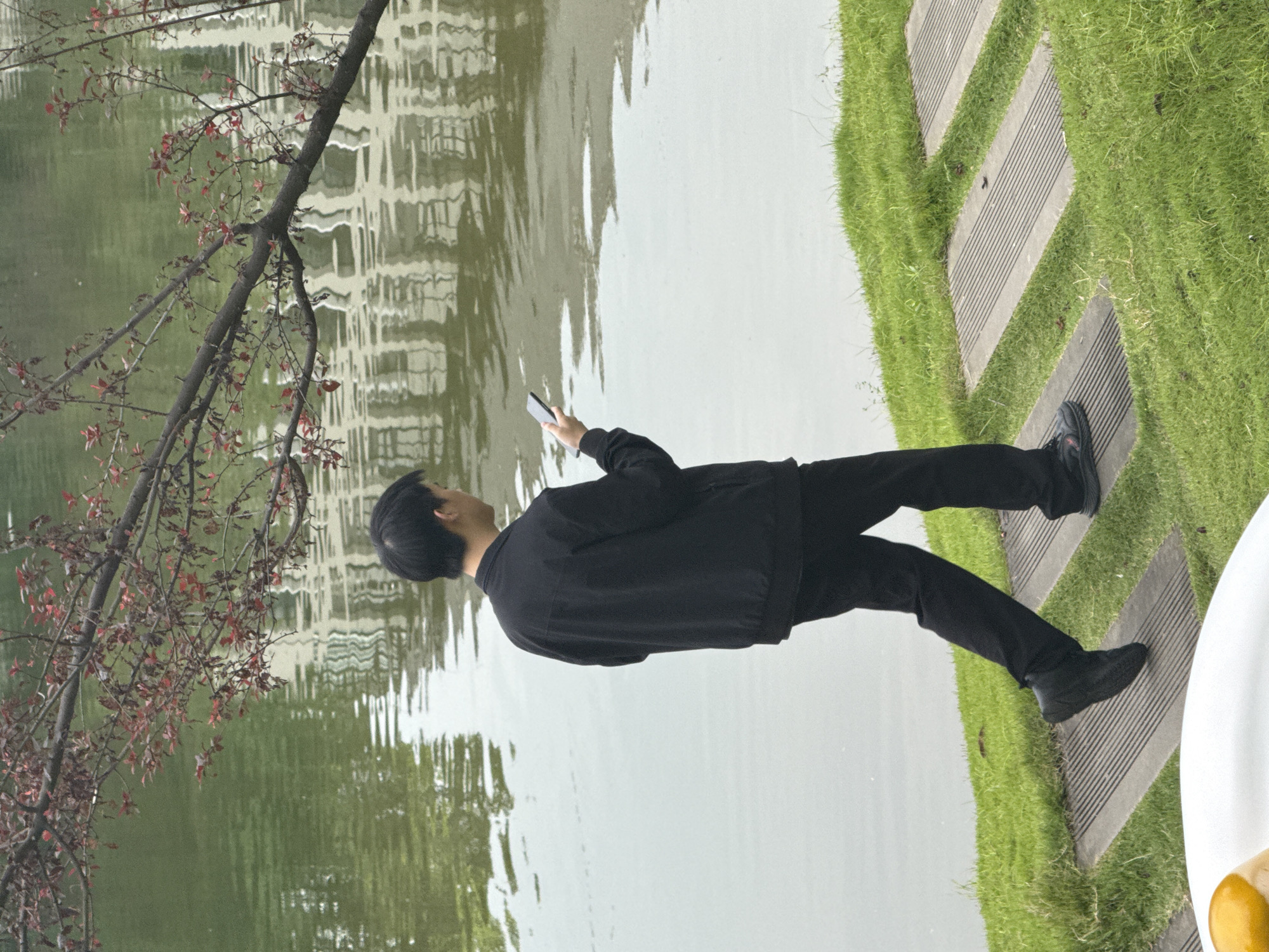 A young East Asian man walks along a paved path beside a body of water. He is wearing a black jacket and black pants, and he is looking at a smartphone in his right hand. The path is bordered by green grass. In the background, there is a tree with red leaves and a white structure reflected in the water. The location is identified as Jiangjin, China.