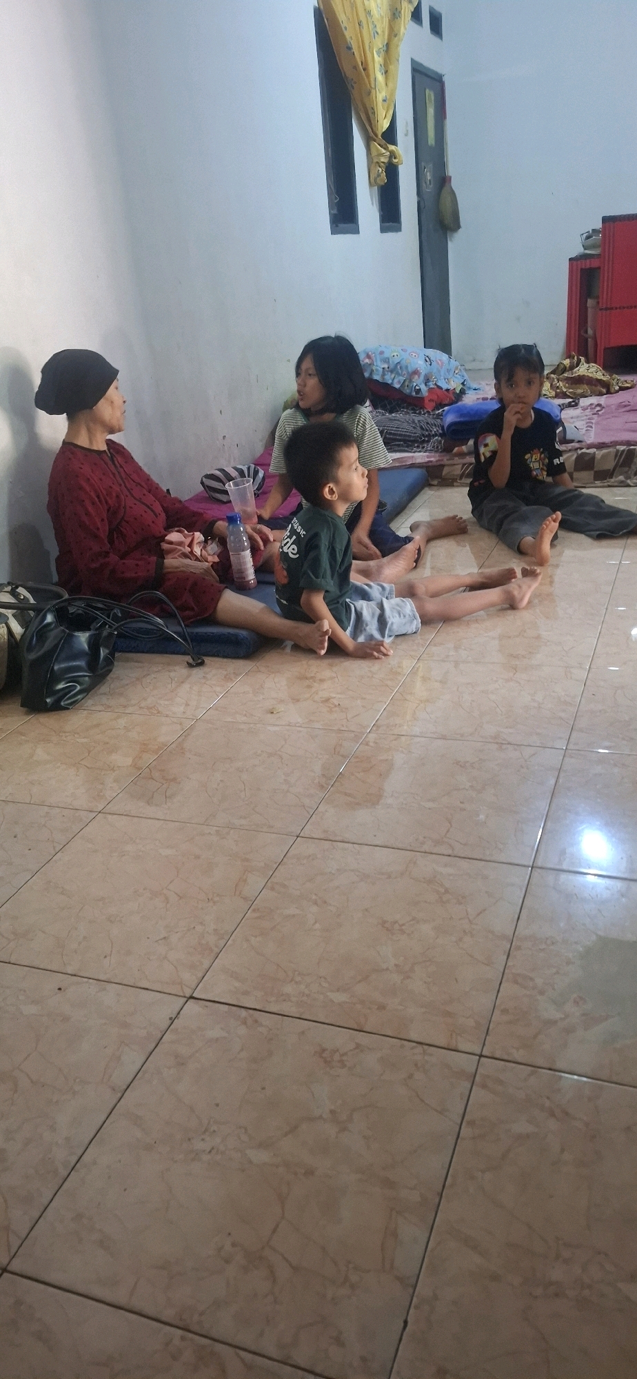 The image depicts an indoor scene, likely a living area or a room in a modest dwelling, situated in Cibinong Hilir, Indonesia. The floor is tiled with a light brown, marble-patterned ceramic. In the foreground, a black leather tote bag rests on a blue mat.
Four individuals are present. On the left, an elderly woman wearing a dark head covering and a maroon long-sleeved shirt with a patterned design is seated on a blue mat. To her right, a young girl with dark, shoulder-length hair is seated, looking towards the left. In front of her, a young boy in a dark green t-shirt and light blue shorts is sitting, looking to his right. Further to the right, another young boy, wearing a black t-shirt with colorful graphics and dark pants, is seated, looking forward and to his left with his hands near his mouth.
Scattered around the individuals are various objects. A clear plastic cup and a bottle of pink liquid are positioned near the elderly woman. Several rolled-up mats and cushions are visible behind the children, suggesting a makeshift sleeping or resting area. In the background, a red cabinet can be seen, and to the left, a portion of a wall with two dark rectangular window frames and a yellow floral curtain is visible. A broom leaning against the wall is also present. The overall lighting suggests it is daytime, possibly late afternoon, with ambient light entering the space.