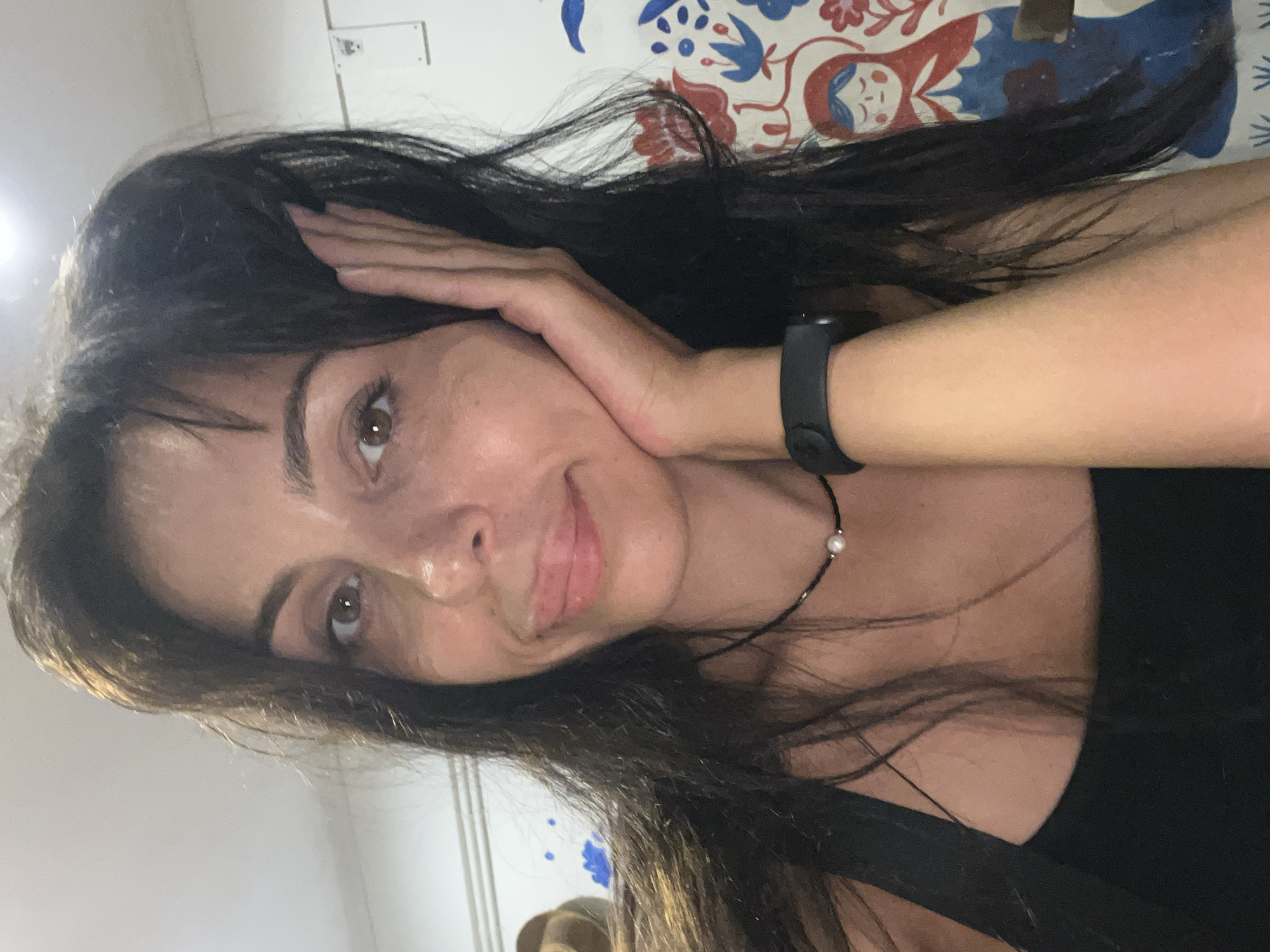 A woman with dark hair and brown eyes is facing the camera, her left hand touching her left cheek. She is wearing a black tank top and a black fitness tracker on her right wrist. A delicate black necklace with a small pearl is visible around her neck. The background includes a patterned surface with red, blue, and white designs, possibly a wall or fabric. The location is listed as Unknown, Indonesia.