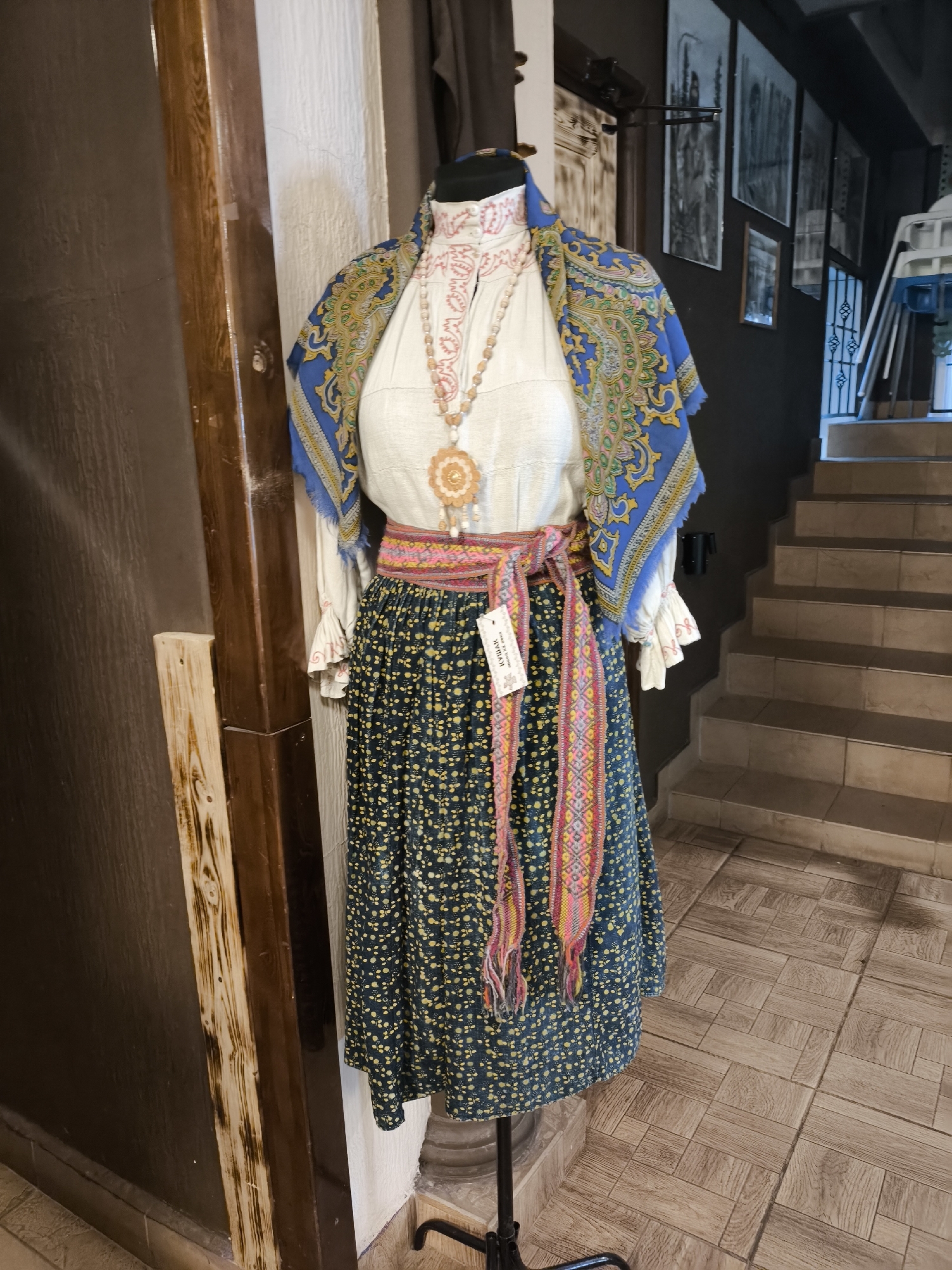 A mannequin displays a traditional Eastern European folk costume in what appears to be a shop or museum in Perm, Russia. The costume consists of a white embroidered blouse with ruffled cuffs, a colorful patterned scarf draped around the shoulders, a wide woven belt with tassels, and a dark blue skirt with a yellow floral pattern. A beaded necklace with a large pendant hangs around the mannequin's neck. The background features wooden elements, framed artwork on a wall, and a staircase. The flooring is tiled in a wood-like pattern. The lighting suggests an indoor environment.