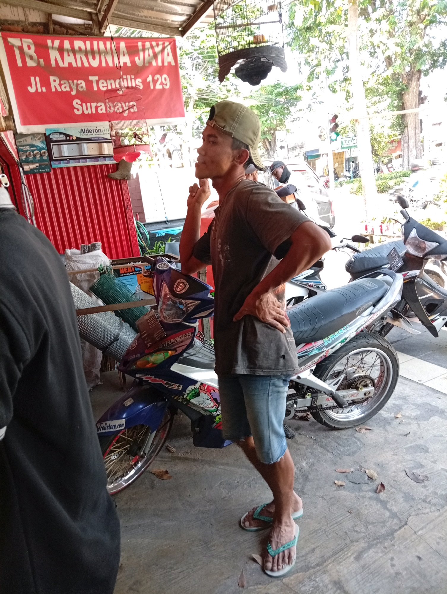 In Surabaya, Indonesia, a man in a green cap and a brown t-shirt, wearing denim shorts and flip-flops, stands beside a blue and white motorcycle. The motorcycle has a license plate that reads "3869 UG". Behind the man, a red banner displays "TB. KARUNIA JAYA" followed by "Jl. Raya Tendilis 129" and "Surabaya". A birdcage hangs in front of the banner, containing a bird. Rolled-up mesh material and other items are visible on shelves to the left of the man. The ground is a concrete surface with scattered leaves. In the background, trees, a traffic light, and other vehicles can be seen.