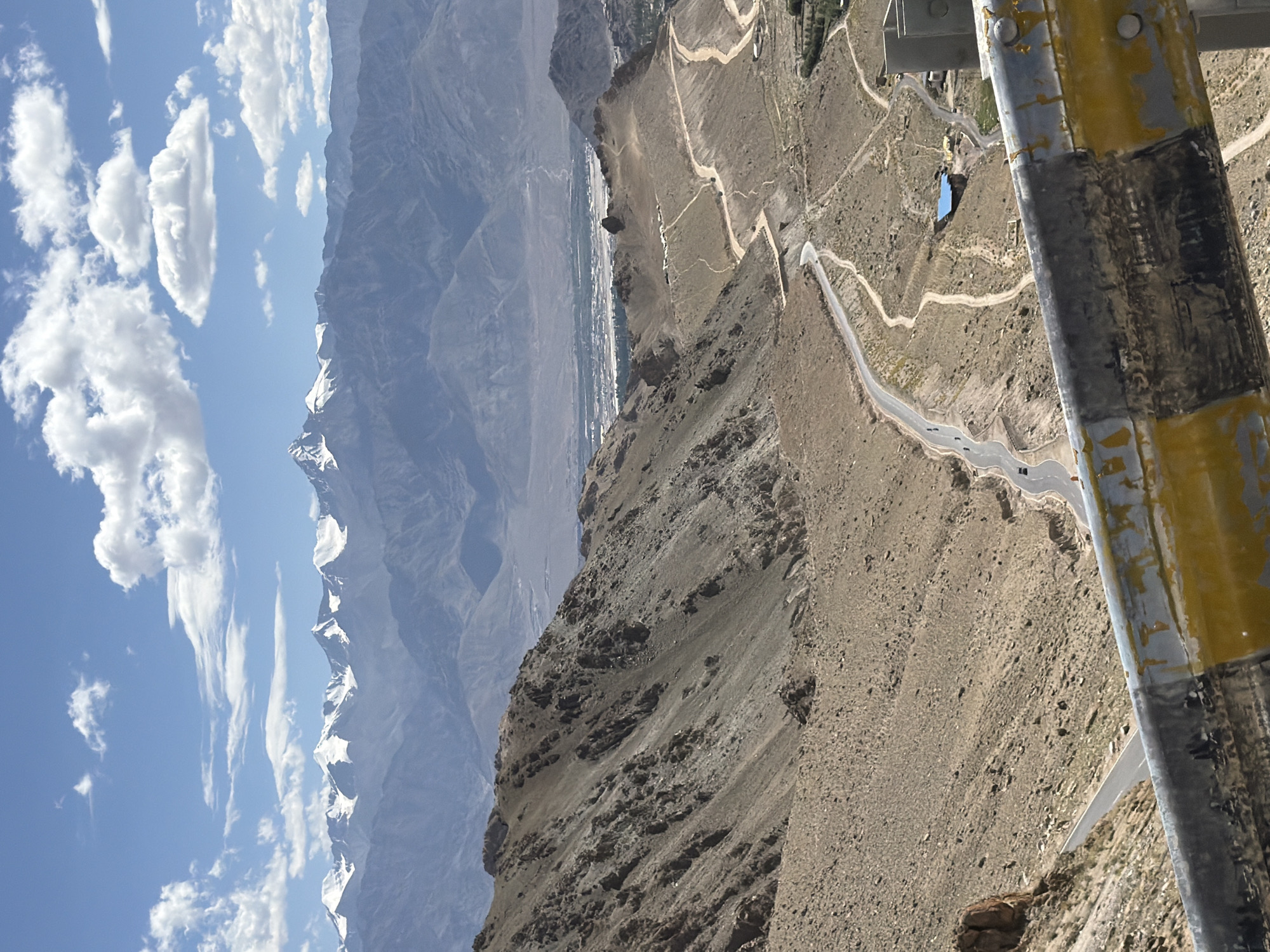 The image captures a panoramic view of the stark, mountainous landscape surrounding Leh, India. In the foreground, a yellow and gray guardrail runs along the edge of a road, with the arid, rocky slopes of a mountain falling away to the left. A winding asphalt road snakes its way down the mountainside, with a few cars visible. Further in the distance, a valley unfolds, dotted with small buildings, including a prominent structure with a blue roof. Beyond the valley, a range of majestic, snow-capped mountains dominates the horizon, set against a bright blue sky with scattered white clouds. The overall impression is one of vast, rugged beauty and remote wilderness.