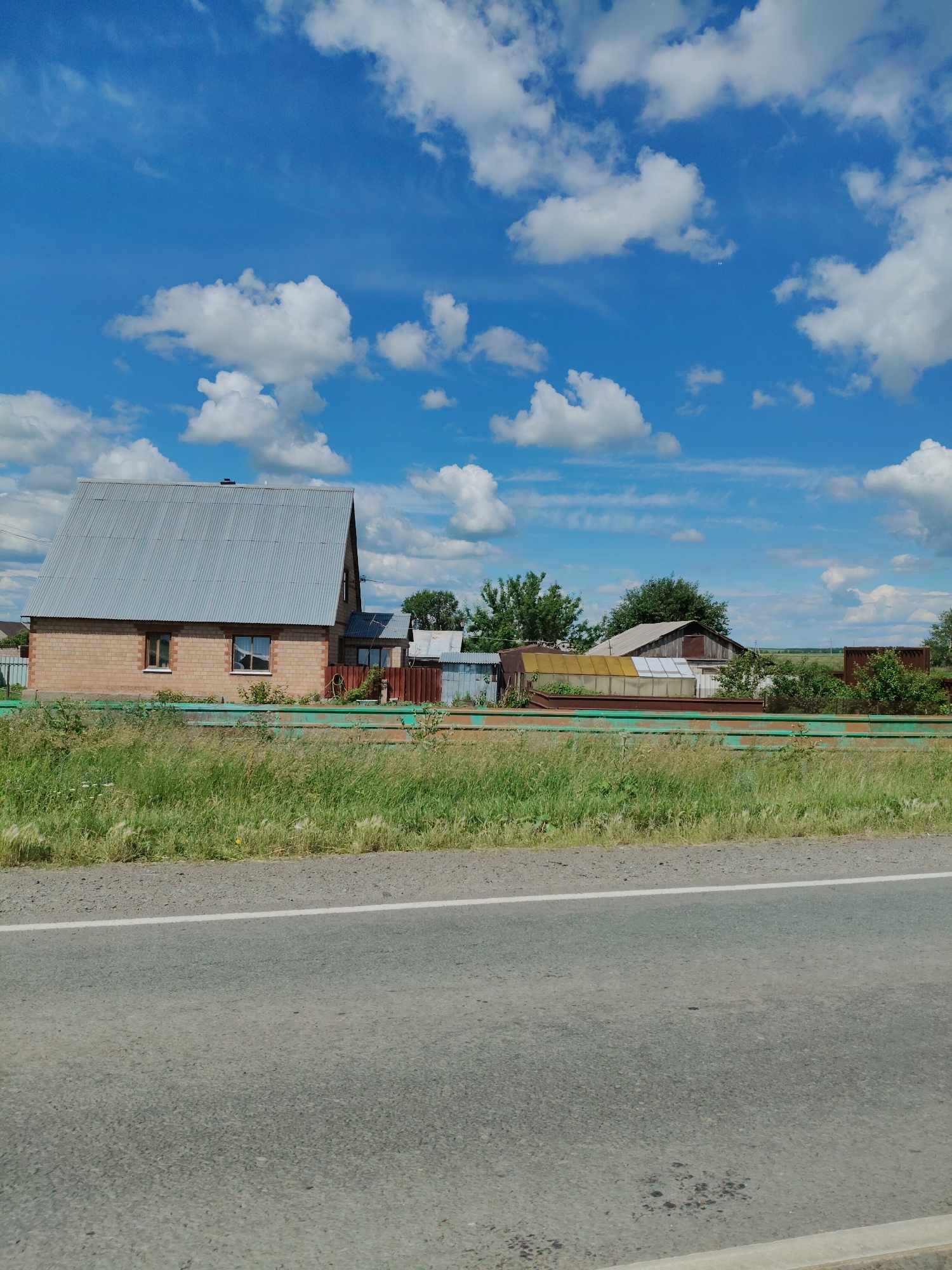 The image captures a rural landscape under a bright blue sky with scattered white clouds. In the mid-ground, a brick house with a corrugated metal roof is visible, flanked by a green fence and tall green grass. Behind the house, several other structures, including sheds and what appears to be a greenhouse, are present. A paved road with a white stripe runs along the bottom of the frame, suggesting the scene is viewed from a roadside perspective. The environment appears to be consistent with a village or countryside setting. The location is specified as Ozerne, Russia. There are no people or animals depicted in the image.