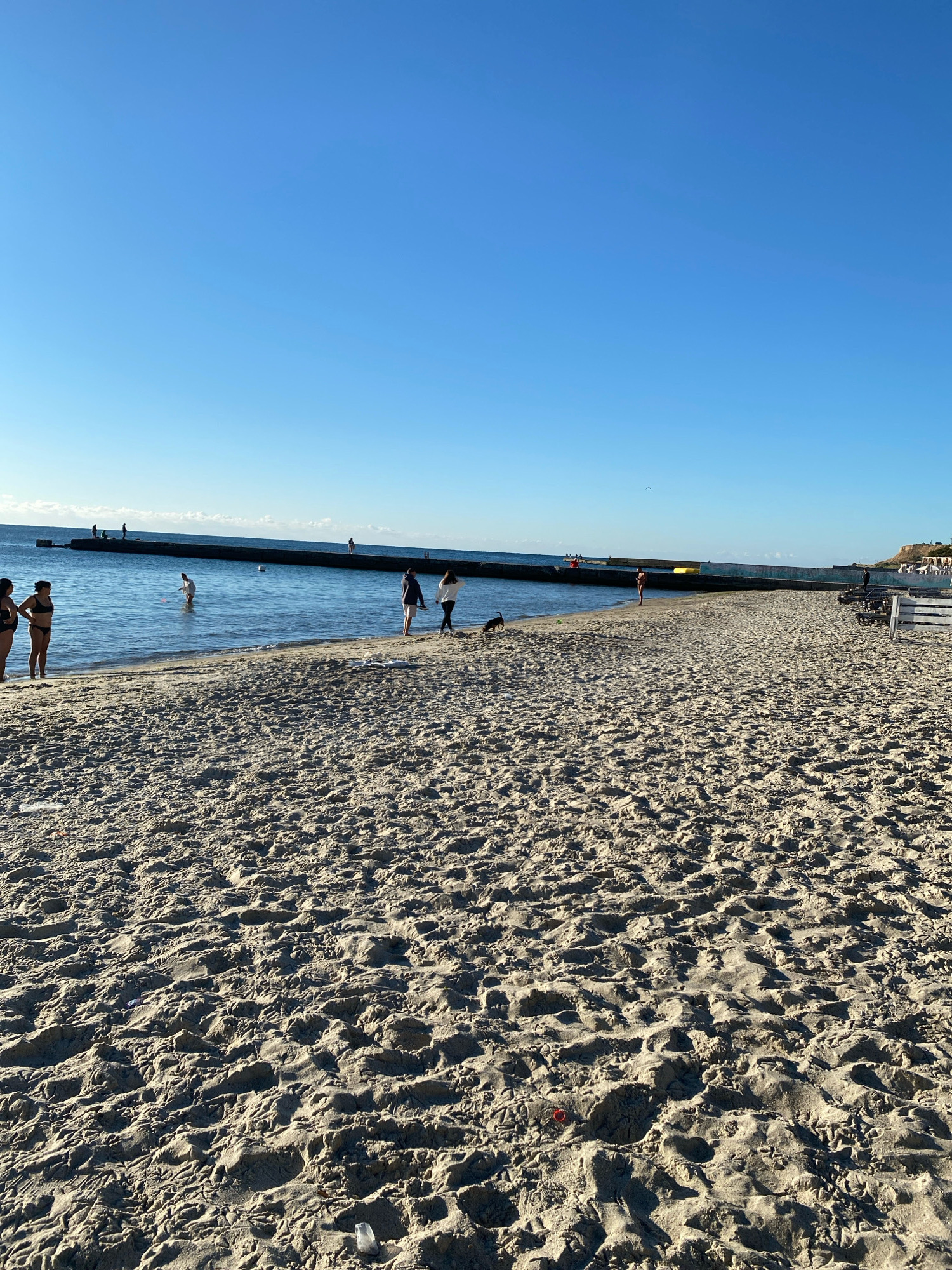 A wide shot shows a sandy beach in Odesa, Ukraine, with the Black Sea in the background under a clear blue sky. Several people are scattered across the beach and in the shallow water. On the left, three women are standing near the water's edge, two in swimwear. Further down the beach, two women are walking, one with a dog. A person in swimwear is standing in the water. In the distance, a long concrete pier extends into the sea, with several figures visible on it. The sand is textured with footprints and small debris.