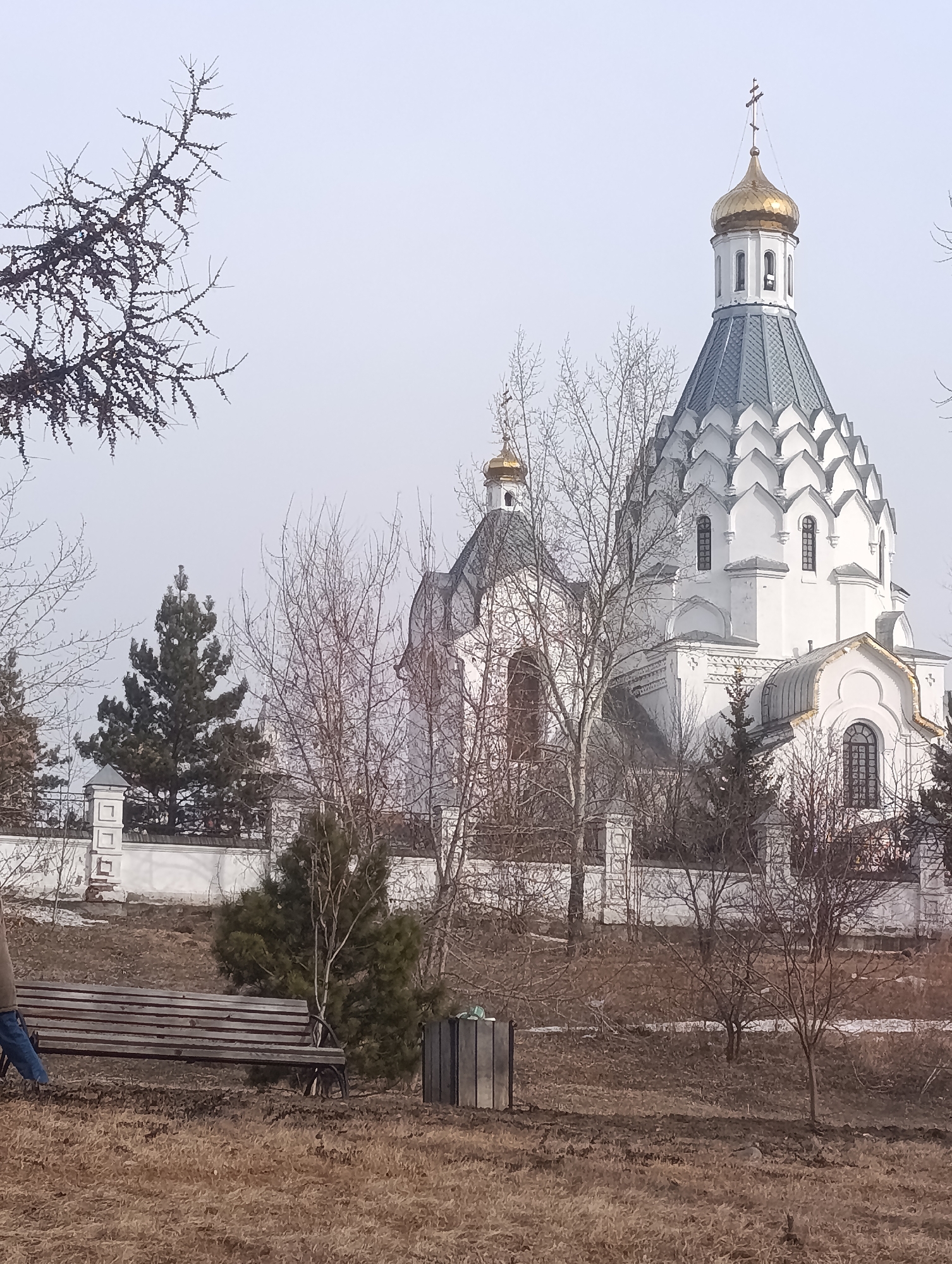 The image captures the Annunciation Cathedral in Krasnoyarsk, Russia. The cathedral, distinguished by its white walls, gray roof, and golden domes, stands behind a low white fence. In the foreground, a wooden bench sits on a patch of dry grass and dirt. Several leafless trees surround the bench and cathedral. A person's leg is visible on the left side of the bench. The sky is overcast.