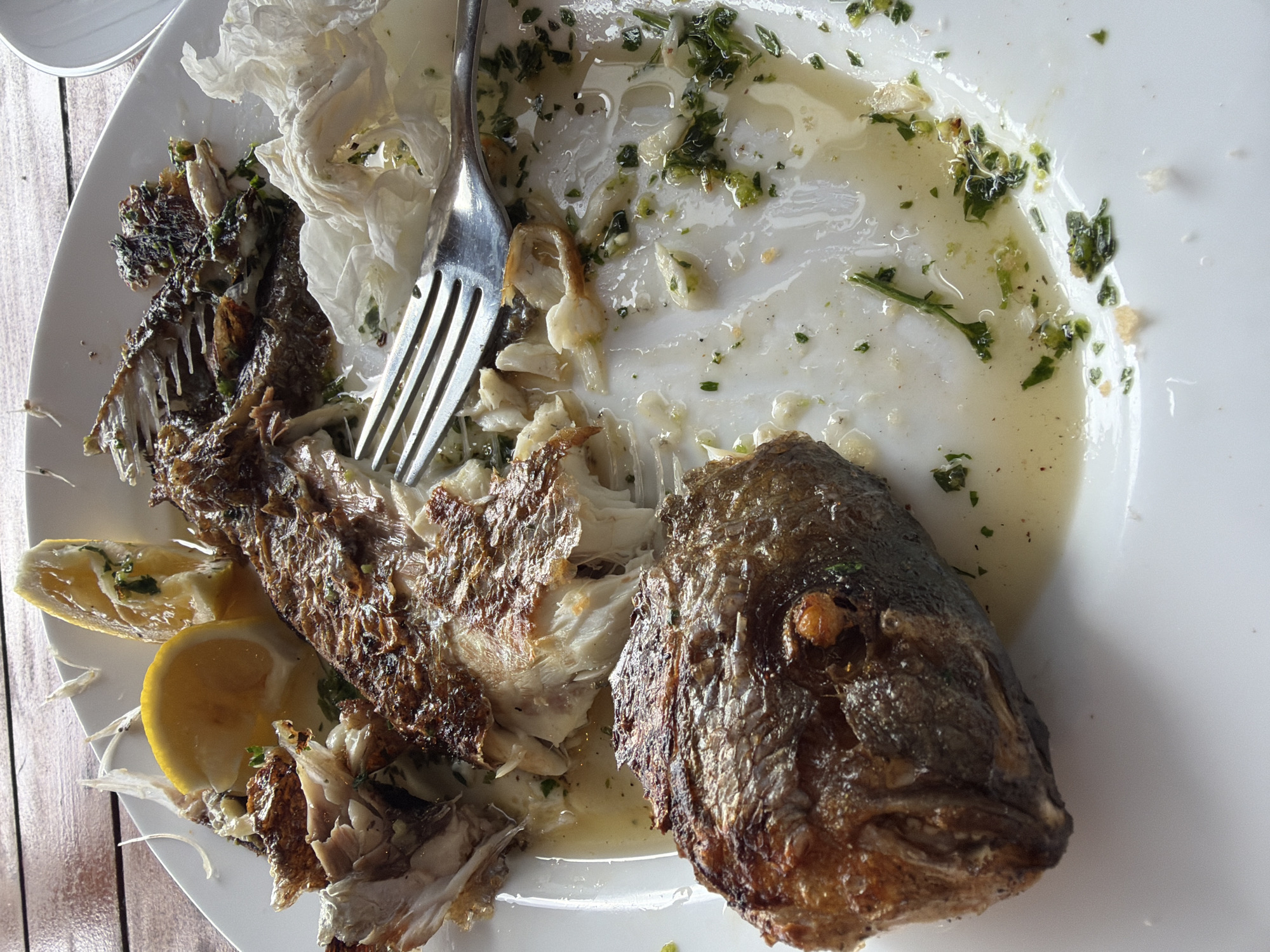 An overhead shot shows a partially eaten fish dish on a white plate, resting on a wooden surface. The fish head sits on the right side of the plate. A silver fork is entangled in the remaining fish parts and a crumpled white napkin sits behind the fork. Two lemon wedges lie near the fish remains. Green herbs and a light-colored sauce cover the plate. The image captures a meal at an outdoor restaurant, potentially located in Donji Orahovac, Montenegro.