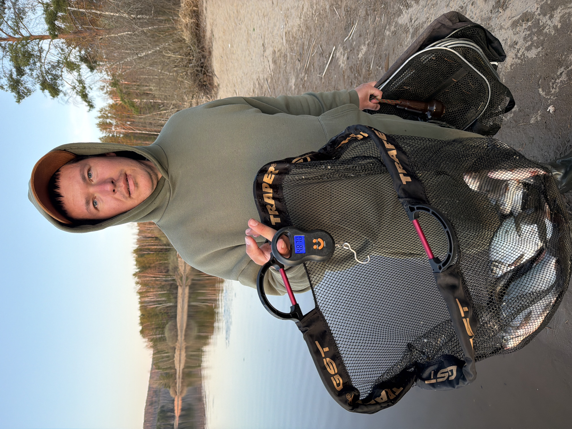 A man stands outdoors holding fishing equipment near a body of water. He is wearing a hooded sweatshirt. He is weighing a net full of fish. He is holding an electronic scale with a digital readout of "4.870". Two fishing nets are visible. One net appears to be full of fish. The background includes trees, land, and the sky. The location is in the city of Chaykovsky, Russia.