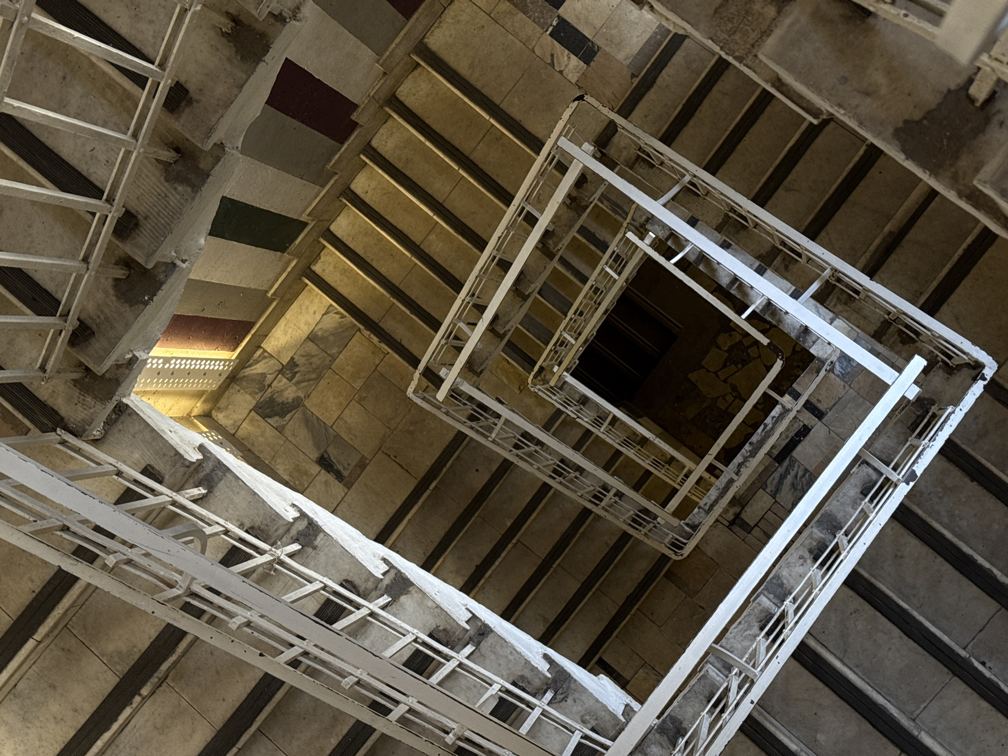 Looking directly down into a square, multi-level staircase, possibly located in Moscow, Russia. The stairs appear to be made of stone or concrete. The railings are white and square. The floors at the very top appear to be decorated with black and red tiles. Each stair has a dark brown, rubber like grip.