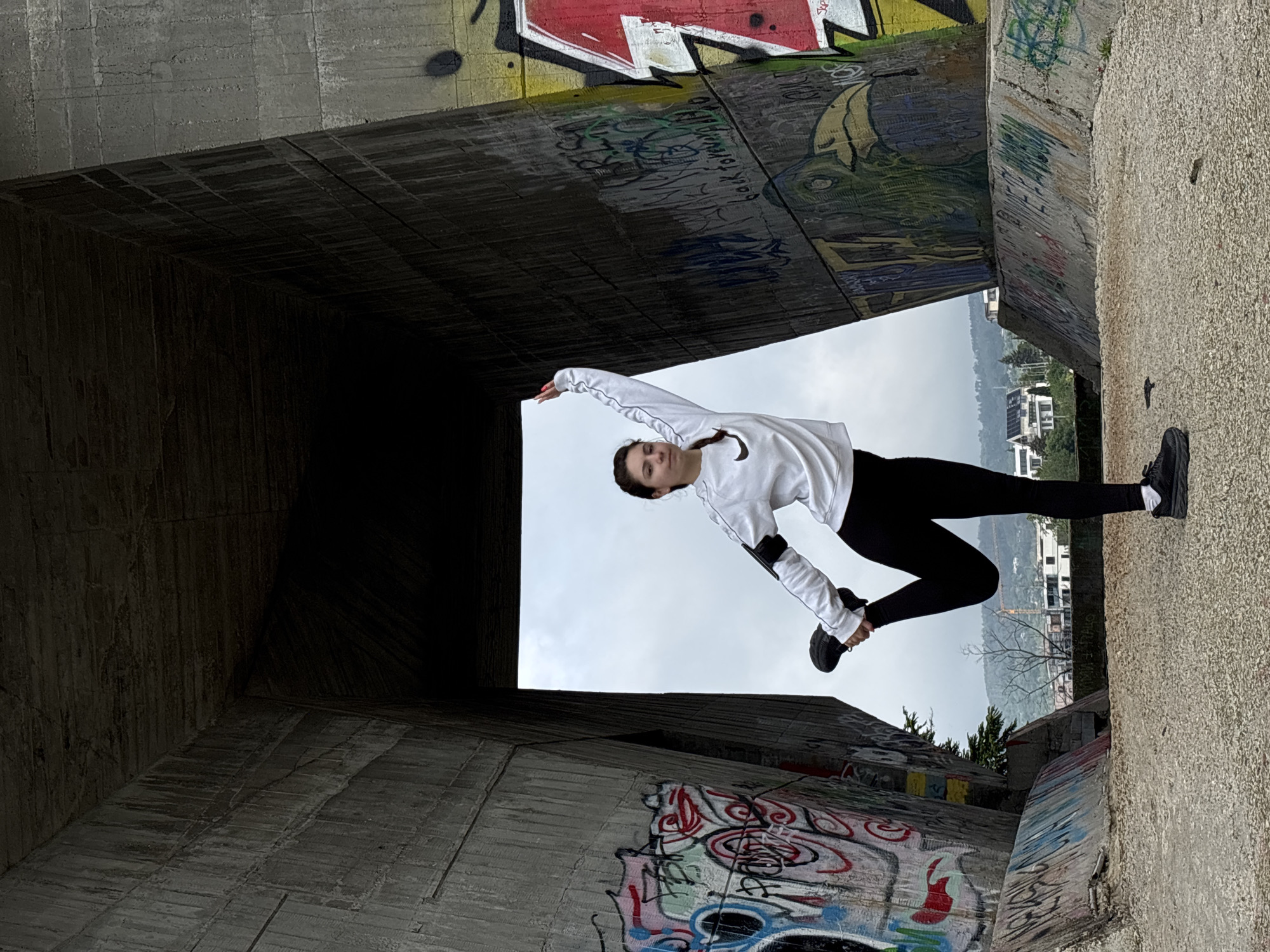 The image shows a young woman in black leggings and a white sweatshirt striking a dynamic pose within a concrete structure adorned with graffiti. The structure appears to be an underpass or a section of a bridge. The background visible through the opening reveals a cityscape with buildings and possibly a hillside, suggesting an urban environment. The photo is taken from an unconventional angle, making the scene appear tilted. The location is likely Varna, Bulgaria.
