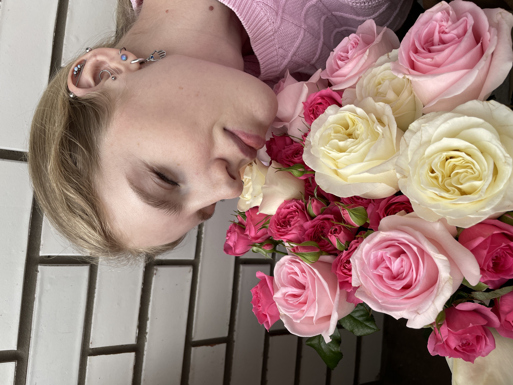 A close-up, high-angle shot captures a person lying on their side with their eyes gently closed, positioned alongside a vibrant bouquet of roses. The subject is a young individual with fair skin and light brown or blonde hair, visible at the top and left of the frame. They are wearing a pink, cable-knit sweater. Their left ear is adorned with numerous piercings, including an industrial bar, helix, conch, and tragus piercings, featuring small opalescent studs and a Hamsa hand charm dangling from an upper cartilage piercing.
The bouquet of roses features a mix of creamy white, pale yellow, light pink, and vibrant fuchsia blooms, ranging from tight buds to fully open flowers. The roses are densely packed, creating a lush and soft backdrop against the person's face. The person's lips are slightly parted, and their expression appears serene and peaceful.
The background consists of a white tiled surface, possibly a wall or floor, with dark grout lines creating a grid pattern. The lighting suggests an indoor setting. There is no visible text in the image. The scene evokes a sense of tranquility and beauty, likely a posed portrait taken in Izhevsk, Russia.