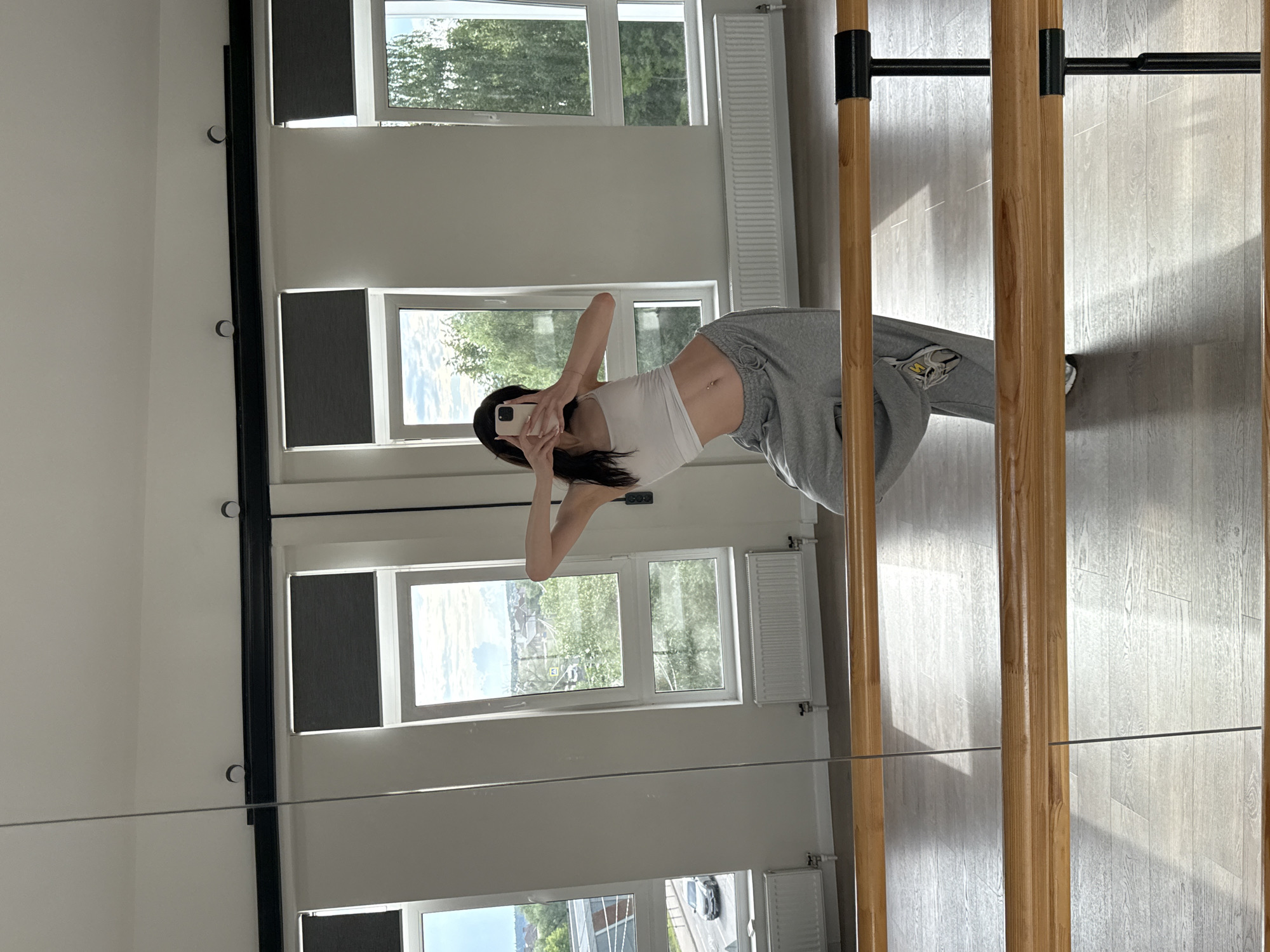 A young woman is captured in a mirror selfie within a dance studio, located in the city of Zakharovo, Russia. She is wearing a white crop top and grey sweatpants, with a phone held up to take the picture. Her pose is dynamic, with one leg bent and arms raised. The studio has large windows offering a view of the surrounding cityscape and trees. The floor is wooden, and a ballet barre runs across the foreground of the image. The room is brightly lit by natural light streaming through the windows.