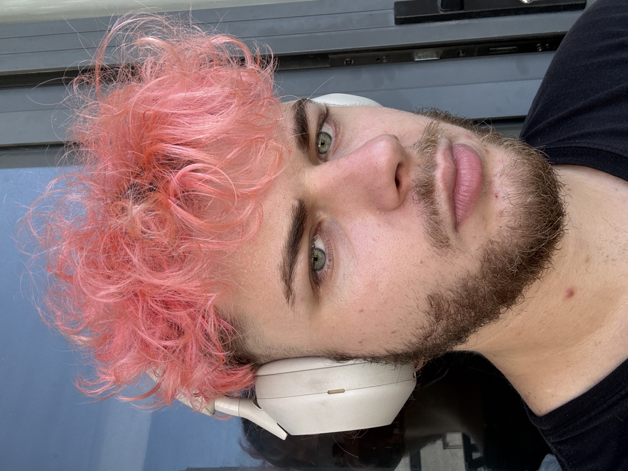 A close-up, selfie-style shot of a young adult male with light skin, facial hair, and pink hair. He is wearing white headphones around his neck. He has light green eyes. He is wearing a black shirt. The background includes an exterior surface, possibly a window sill, reflecting a blue sky. There is no indication of the location being Đà Nẵng, Vietnam, and no specific urban details are present.