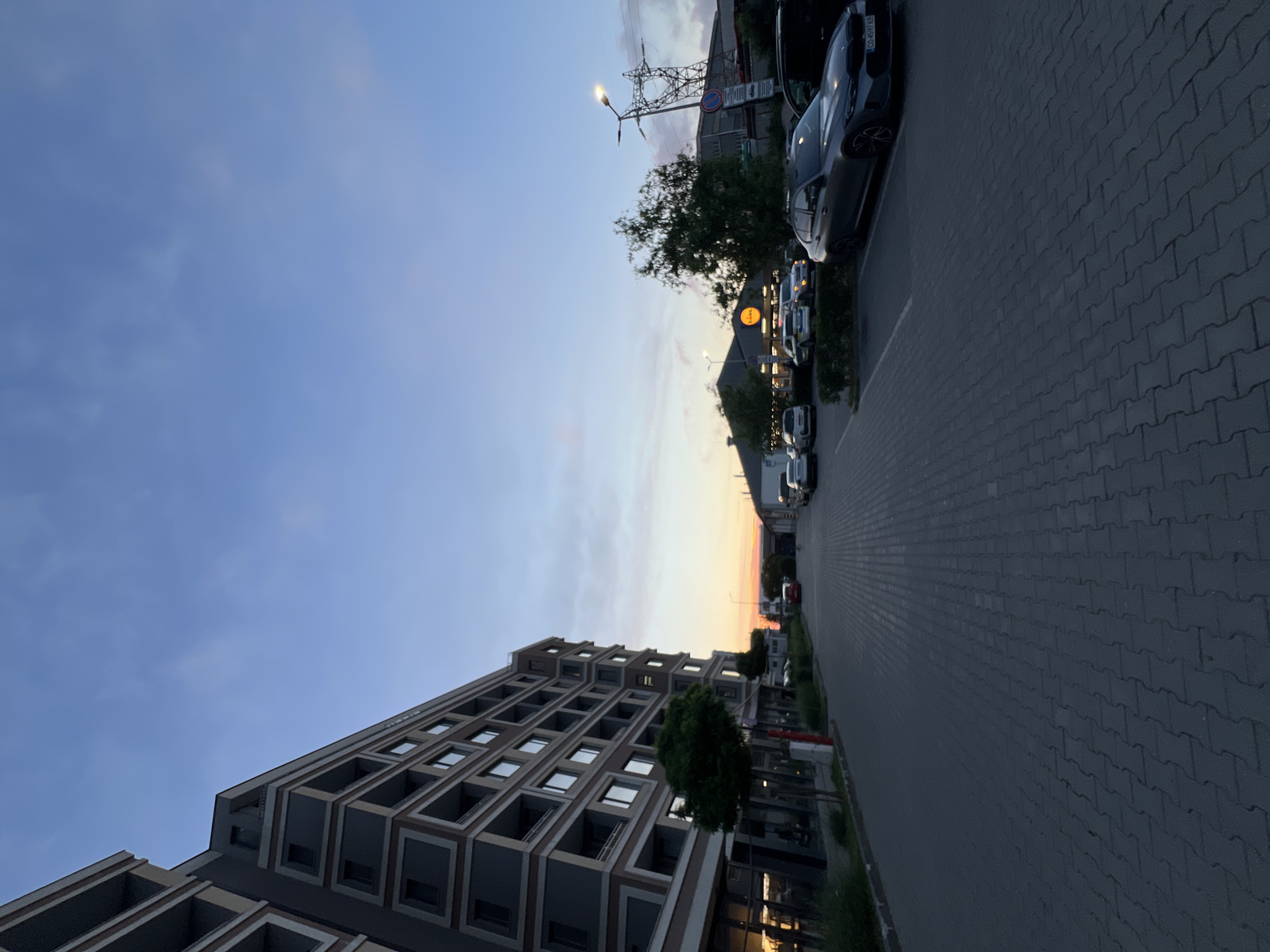 The image depicts an outdoor scene in Burgas, Bulgaria, at dusk. On the left side of the frame, a modern, multi-story building with a grey facade and balconies stands prominently. A tree is situated in front of the building. The ground in front of the building is paved with grey interlocking bricks. A street or pathway extends from the foreground into the mid-ground.
In the mid-ground, several cars are parked on the right side of the pathway. The building directly opposite the apartment complex appears to be a commercial establishment with yellow signage, possibly a supermarket. Further in the background, a power transmission tower is visible against the twilight sky. The sky itself transitions from a pale orange glow near the horizon to a deep blue overhead, with scattered clouds. The overall atmosphere is one of quiet evening in a mixed-use urban area.