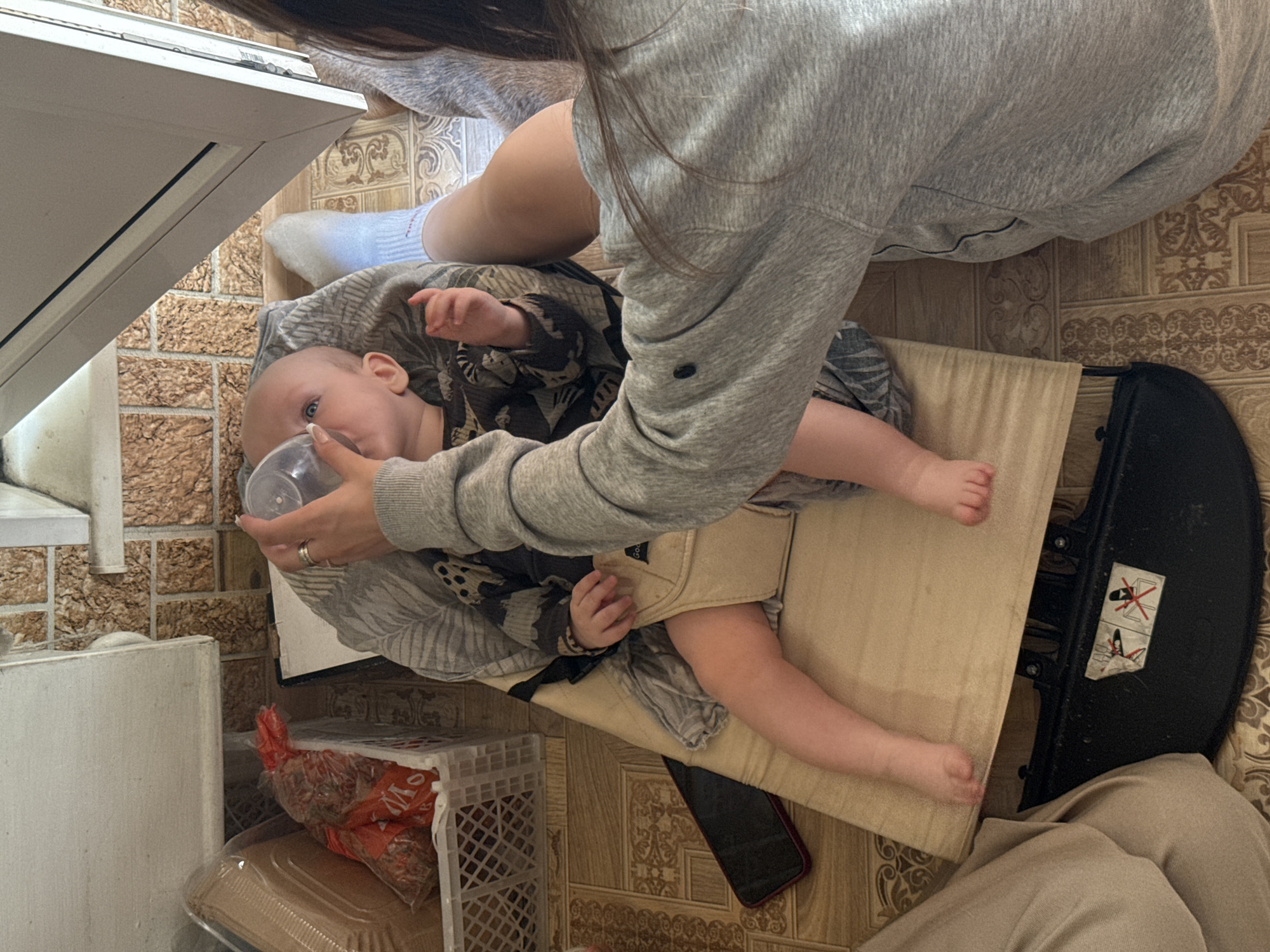 The image captures a nurturing indoor scene, likely within a home in Moscow, Russia. An adult, primarily visible from the chest down, is seated or kneeling on a patterned tiled floor, gently feeding a baby from a clear plastic bottle. The adult wears a gray hooded sweatshirt, light-colored pants, and light blue socks.
The infant, with light hair and blue eyes, is resting comfortably in a beige fabric baby carrier or bouncer with a black frame. The baby is dressed in a patterned, possibly camouflage-style, outfit and appears calm while feeding.
The surrounding environment features a decorative tiled floor and, to the left, part of a white appliance, possibly a radiator or a cabinet. A white plastic crate holds a red mesh bag with partial text "V О" visible, likely indicating its contents. A black smartphone with a red case is lying on the floor beside the baby carrier. On the black frame of the baby carrier, a safety warning sticker with crossed-out symbols (indicating prohibitions like standing or lifting by specific parts) is present. Faintly visible text "GOO" can be seen on the beige fabric of the carrier. The scene conveys a quiet, attentive moment between the caregiver and the infant.