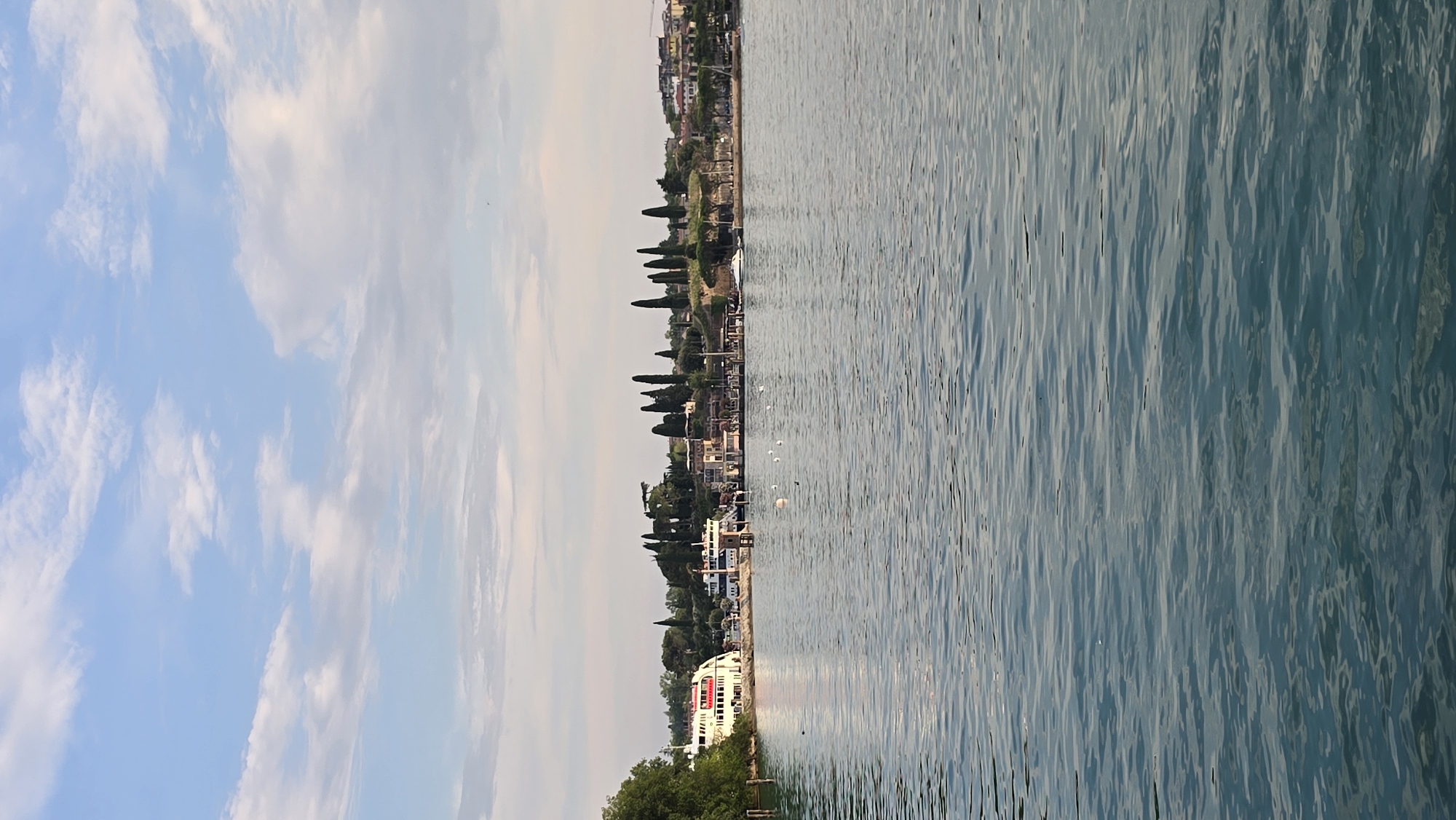 The media file shows a scenic view of a waterfront with a town nestled along the shore and a body of water in the foreground. The scene is oriented vertically, with the sky occupying the left side and the water the right. The sky is a light blue with scattered white clouds, suggesting a clear day.
The waterfront is lined with buildings, trees, and cypress trees, creating a picturesque backdrop. Several boats are visible, including a large white ferry with red accents docked near the shore. White buoys are also scattered on the water. The town appears to be a charming European village, possibly a tourist destination given the presence of a ferry.
The water is a deep blue-green, with gentle ripples reflecting the light. The overall impression is one of a peaceful, sunny day in a Mediterranean setting, likely in Peschiera del Garda, Italy, as indicated by the location context. No people are clearly visible, nor is there any discernable text. The time of day appears to be late afternoon or early evening, judging by the soft lighting.