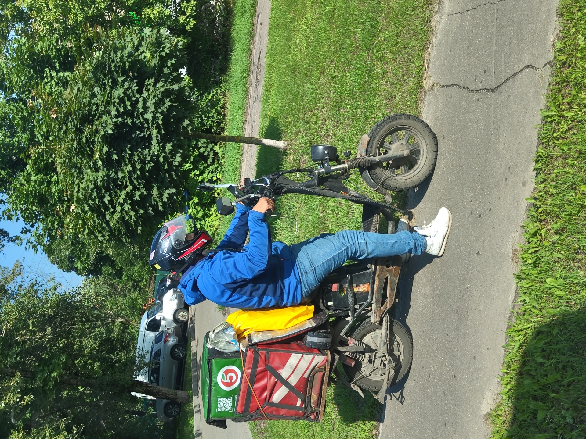 A person wearing a blue hoodie and jeans rides an electric scooter on a paved path in Zhukovsky, Russia. The scooter has a large, red and green insulated delivery box on the back, with a yellow bag tucked behind it. The person is wearing a black helmet with red and white accents. The background features lush green trees and grass, with a few cars parked in the distance. The sky is clear and blue.
