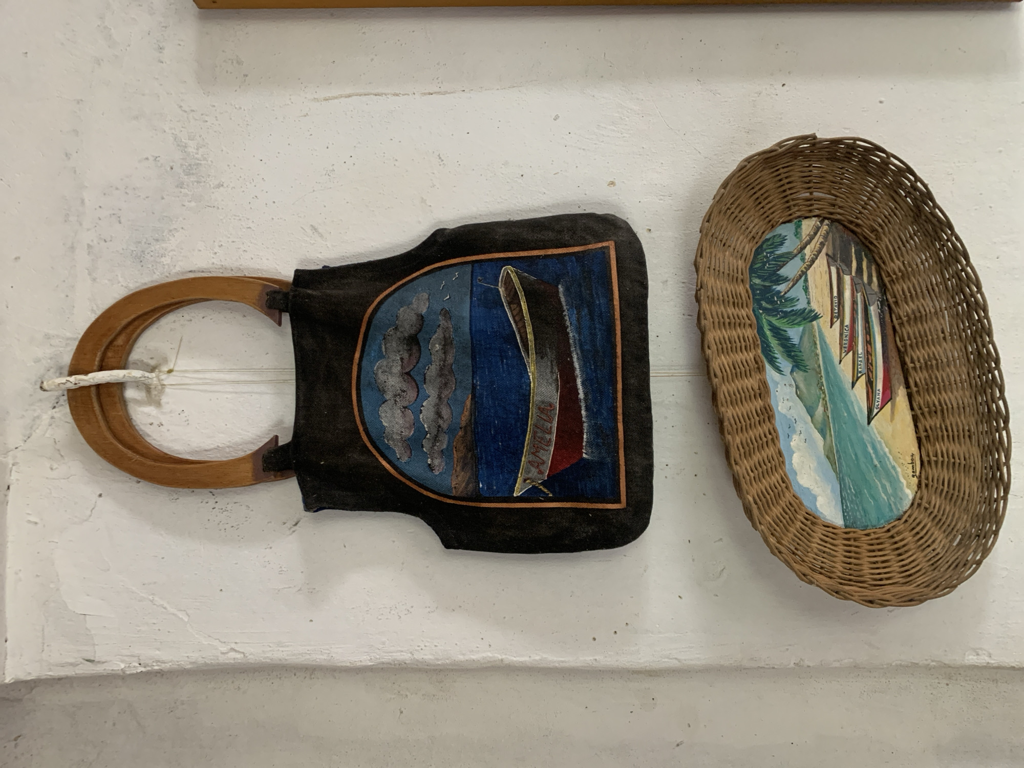 Two decorative items are displayed against a textured, off-white wall, likely within a building in Santa Ana, Venezuela.
On the left is a dark fabric piece, possibly a small bag or wall hanging, with a U-shaped light brown wooden handle. The fabric features a central painting bordered by a thin brown line. The painting depicts a sailing vessel on dark blue water, possibly at dusk or in stormy conditions, with a prominent red and white sail. Behind it, a smaller, older-style sailboat is visible. On the main boat, the name "AMELIA" is clearly legible, and below it, the word "CRUISE" is partially visible. Abstract, cloud-like formations appear on the left side of the painting.
To the right, an oval-shaped, light brown woven wicker basket serves as a frame for a vibrant painting. This artwork portrays a tropical beach scene with lush green palm trees on the left, and turquoise water with white foamy waves on the right. Several colorful boats, possibly traditional canoes or fishing vessels, are depicted on the shore or in very shallow water. Names are visible on some of these boats: "OLYANA" and "VERONICA" are discernable on one boat, and "CRUISE" is partially visible on another. A signature, possibly "gamboao" or similar, is located in the bottom right corner of this painting.
The wall itself shows signs of age, with uneven texture and some minor chipping. The items are illuminated by what appears to be ambient indoor light.