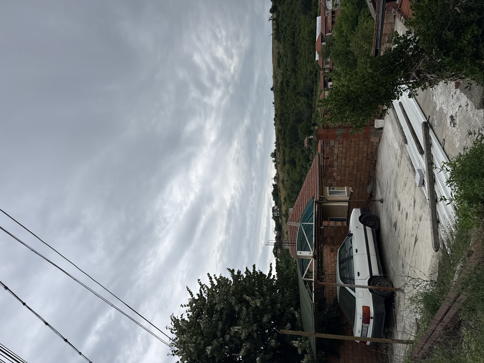 The image captures an overcast day in a rural setting, likely within the city of Slavyani, Bulgaria. The foreground features a white Audi sedan parked under a makeshift shelter constructed with a green corrugated plastic roof supported by metal poles. The car is positioned on a concrete surface. To the right of the car, a brick wall partially encloses the area, and beyond it, a green leafy tree is visible. In the mid-ground, red-tiled roofs of houses are scattered across a gentle, tree-covered hillside that stretches towards the horizon. The sky is filled with grey, wispy clouds, suggesting impending rain or a generally cloudy day. Power lines are prominently visible in the upper left portion of the frame, extending diagonally across the sky. The environment appears to be a residential area in a village or small town, characterized by a mix of residential buildings and natural vegetation.