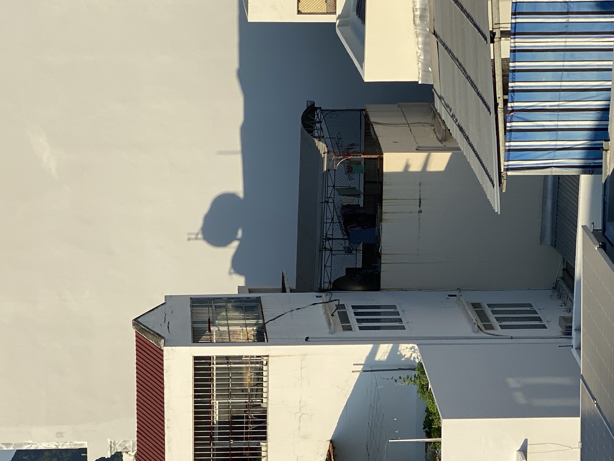 In an urban outdoor scene, the viewer is presented with a partial view of residential buildings under bright sunlight. The foreground features a section of a rooftop with solar panels. To the left, a white building with a red-tiled roof is visible, featuring barred windows on an upper floor and multiple windows on a lower floor. Adjacent to this, another white building has a similar barred window arrangement on its upper section and several windows below. The middle ground shows the rooftop of another building with a covered area, possibly for laundry, displaying clothes hanging on a line. In the background, a large, plain white wall dominates, casting a long shadow from an unseen object, possibly a water tank, which creates a distinct circular silhouette with a handle-like protrusion. The overall environment suggests a densely populated urban area with a focus on everyday living spaces.
