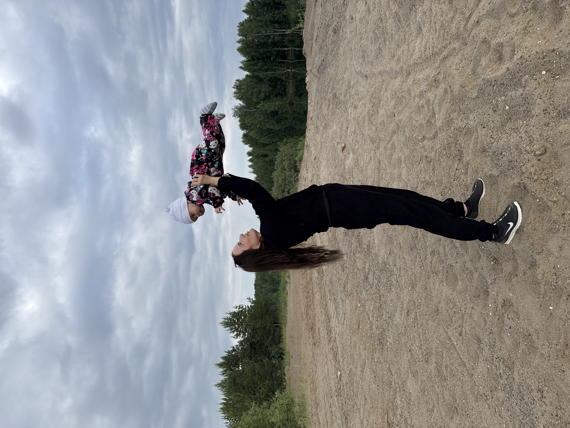 A woman in a black tracksuit and sneakers is standing on a sandy area, holding a baby above her head. The baby is wearing a white hat and a colorful jumpsuit with a Hello Kitty pattern. Behind them is a dense forest and a body of water, possibly a lake or river. The sky is overcast with clouds. The location is described as Novyi, Russia.