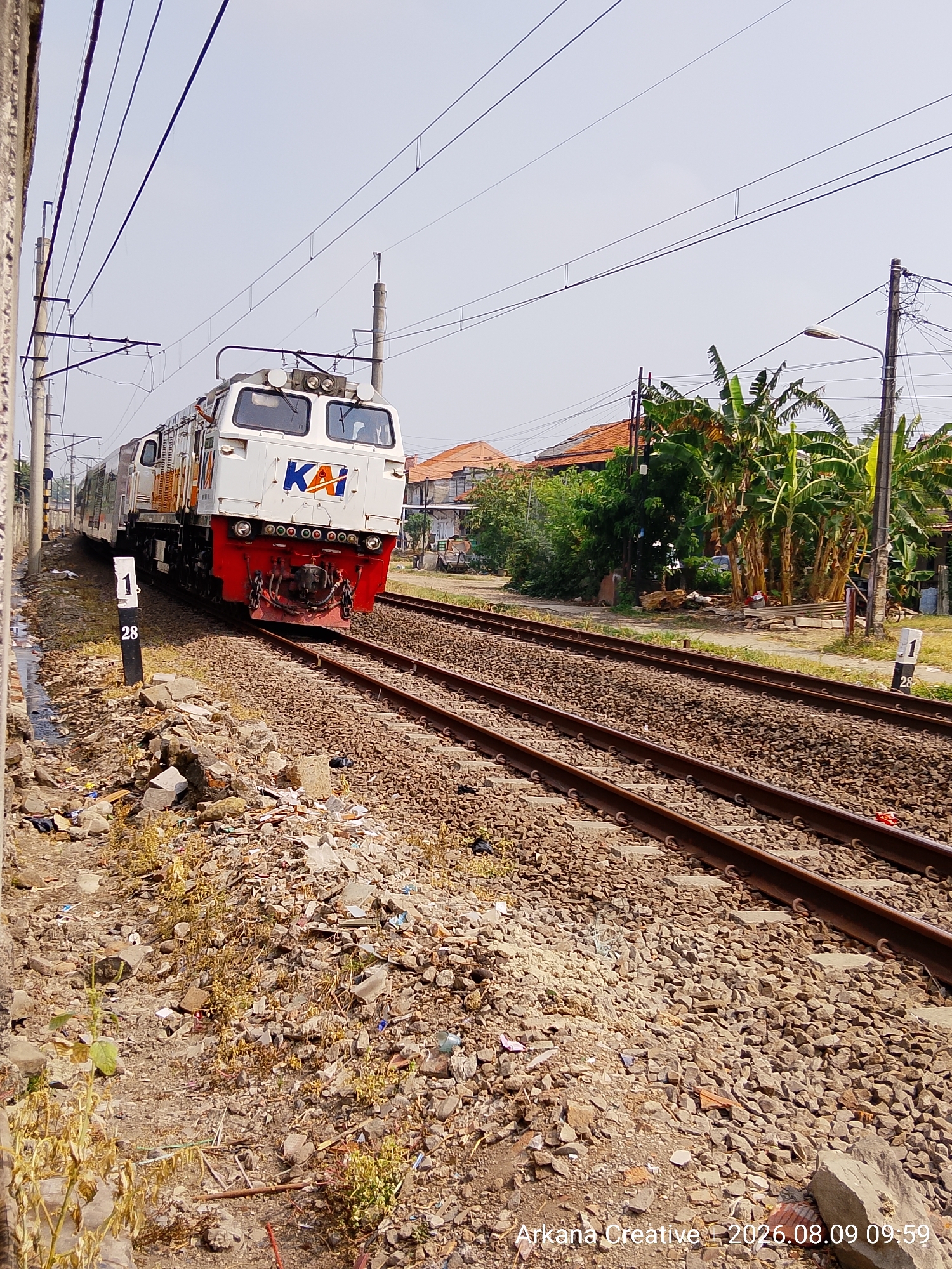 An Indonesian state railway train, identified by the "KAI" logo on its front, is traveling on a railway track in an outdoor, possibly semi-urban or industrial area. The train is predominantly white with red and orange accents on the front. It is moving from left to right in the frame. The railway tracks are well-maintained and covered in ballast. Adjacent to the tracks, on the left side of the image, there is a pile of debris and rubble, including broken concrete, plastic, and vegetation, suggesting an area with construction or waste accumulation. On the right side, there are houses with orange tile roofs, lush green trees, including banana trees, and a utility pole with street lighting. The sky is clear and blue, indicating daylight conditions. A marker post with the number "28" is visible near the tracks on the left. The image is dated "2026.08.09 09:59".