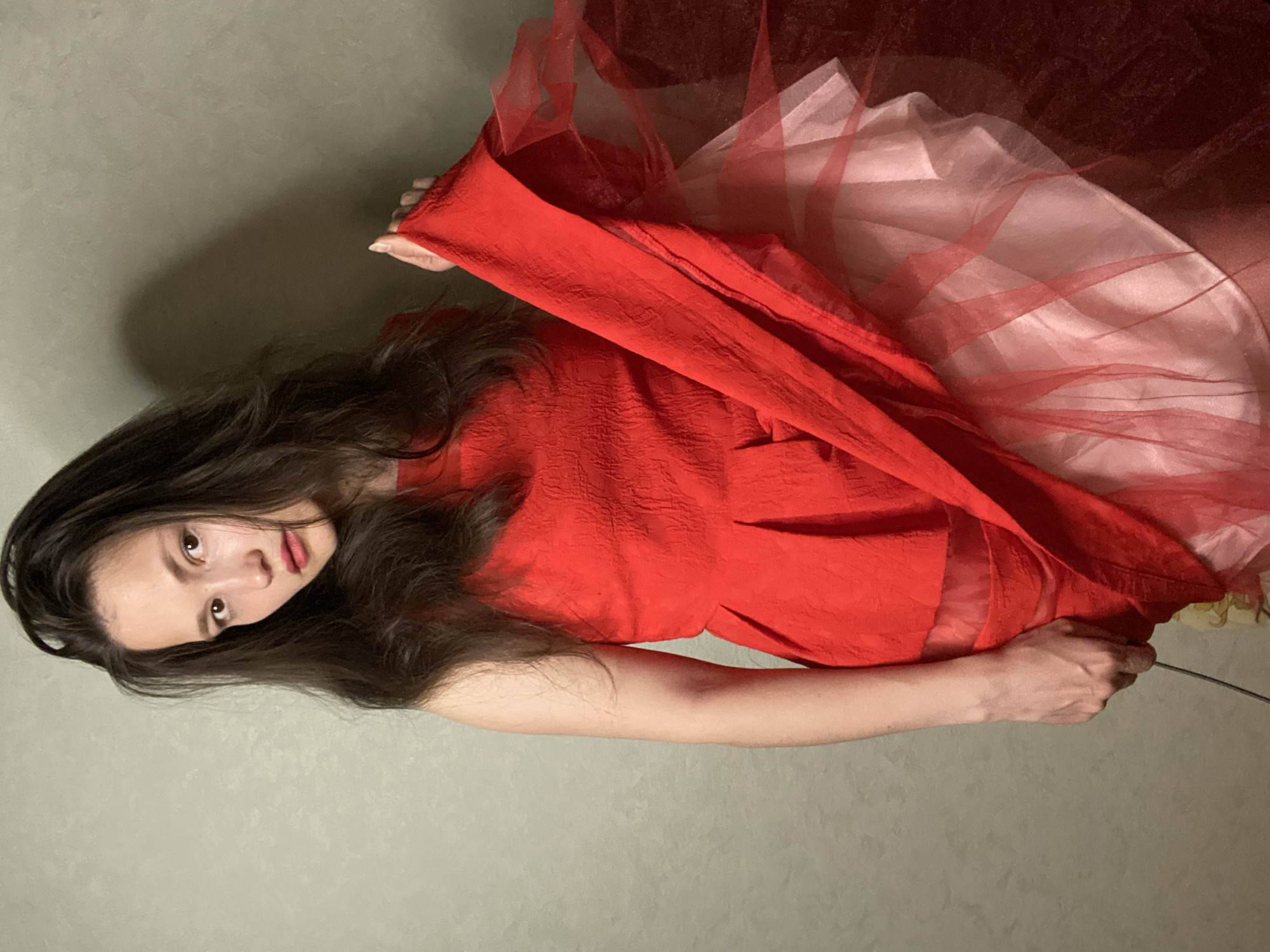 This media file is a portrait of a person wearing a red dress and white and red tulle. The person has long, dark hair and is looking at the camera with a neutral expression. The person's left hand is visible, holding onto the red fabric of the dress. The background is a plain, textured wall in a neutral color. The lighting appears to be artificial, with a shadow cast on the wall behind the person, suggesting an indoor setting. The overall mood of the image is somewhat dramatic due to the contrast of the red dress and the soft lighting. There is no visible text in the image. The location context is Chaikovsky, Russia.