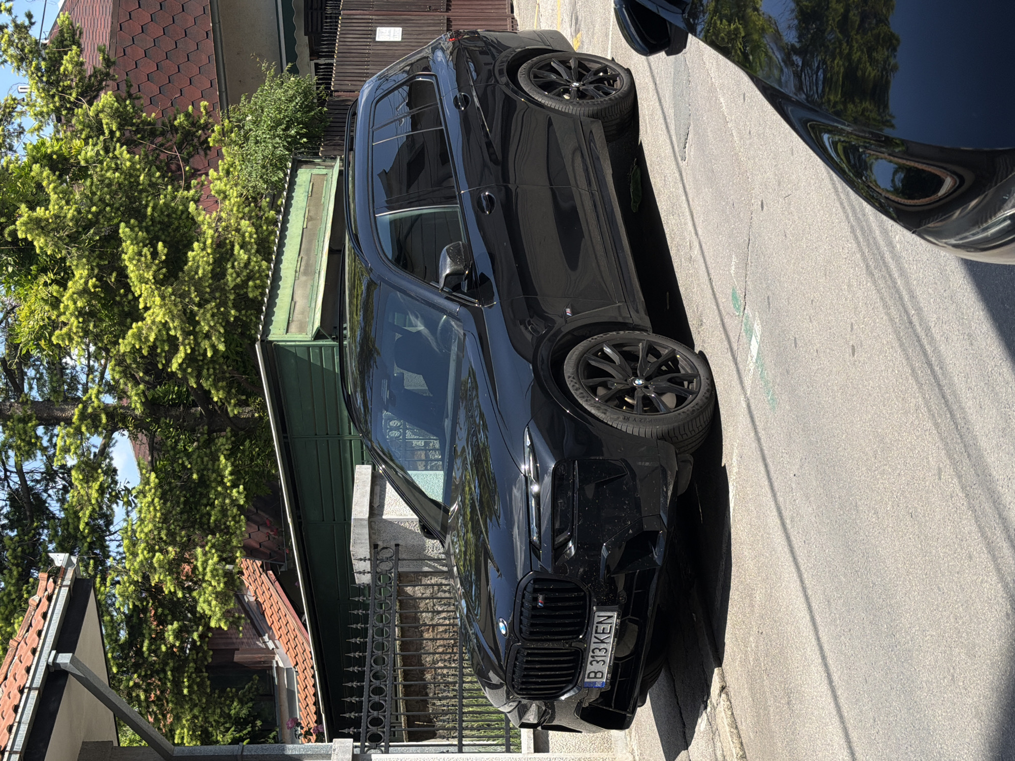 A black BMW X7 M50i is parked on the street. The car has a Romanian license plate B 313 KEN. The environment appears to be a residential area. The location is likely in the city of Belgrade, Serbia. There are houses and trees visible in the background. The photo was taken in daylight. Another car is partially visible on the right side of the image. The car is parked next to a fence and a house with a green roof.