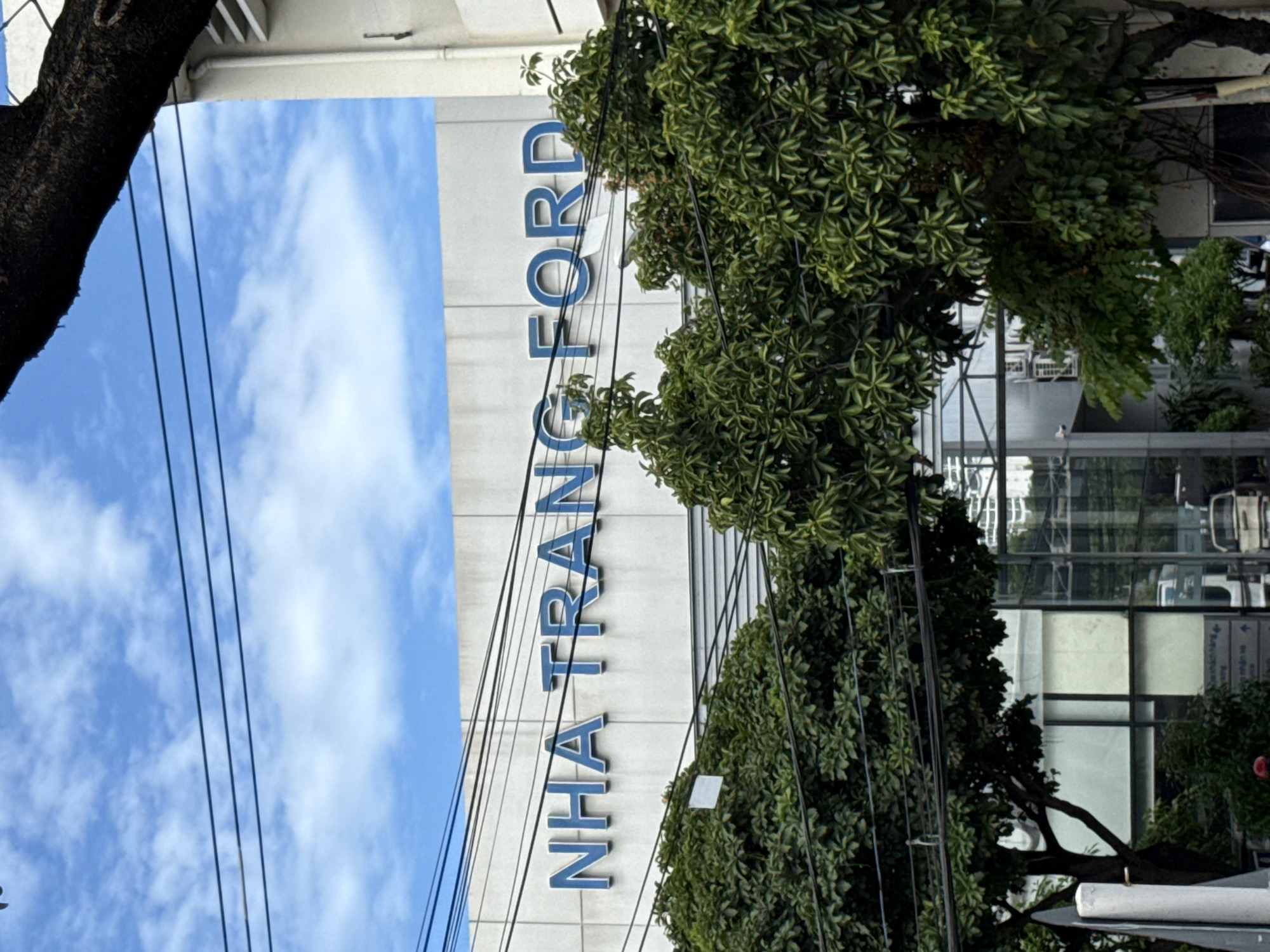 The image shows a building with "NHA TRANG FORD" written in large blue letters across its facade. The building appears to be a car dealership or service center. Lush green trees and foliage are in the foreground, partially obscuring the building. Overhead, power lines crisscross the frame against a partly cloudy blue sky. The architecture suggests a modern commercial establishment. The overall scene depicts a street view in Nha Trang, Vietnam.
