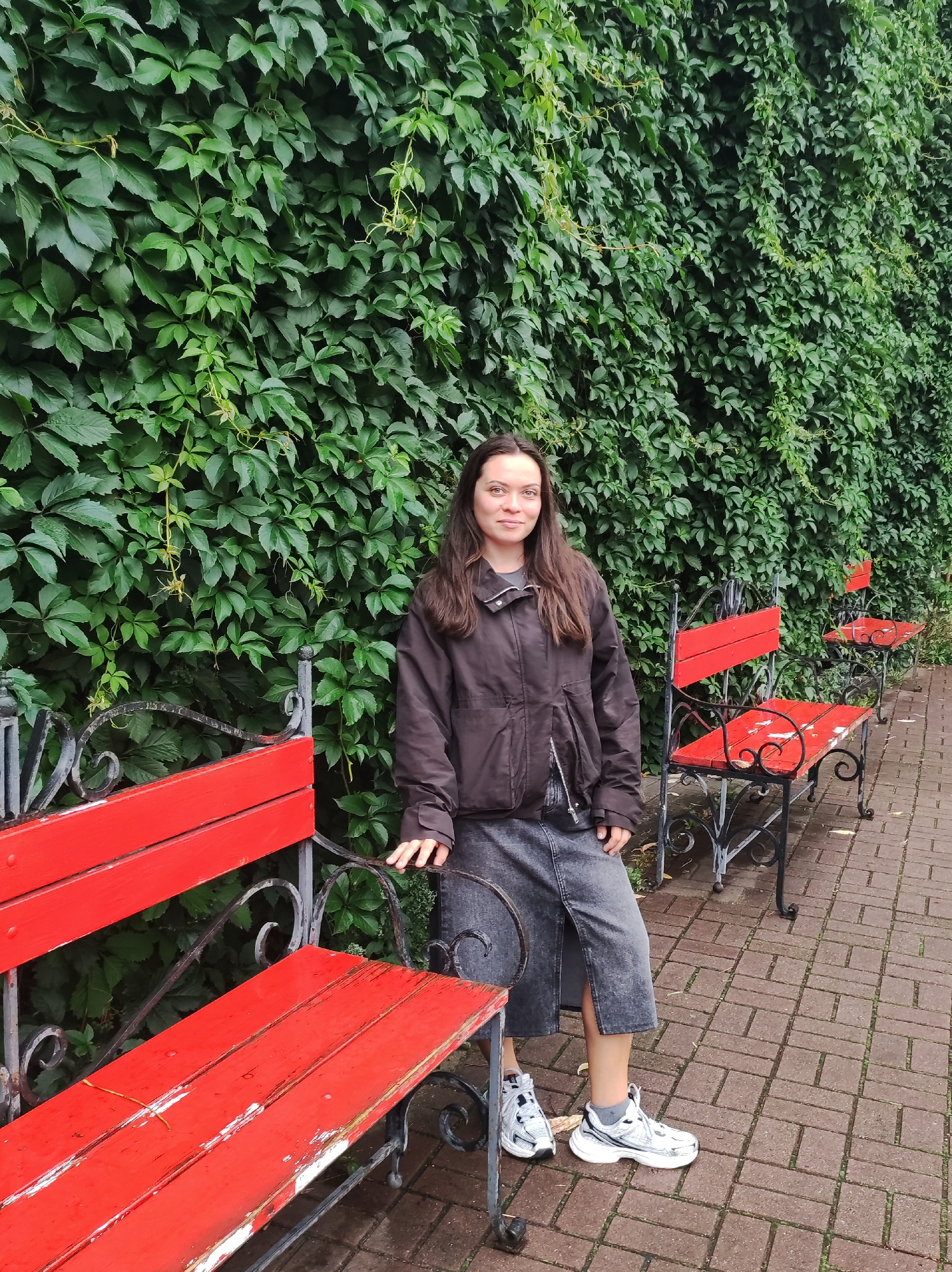 A young woman stands in front of a thick, green ivy-covered wall in Saint Petersburg, Russia. She is wearing a dark brown bomber jacket, a grey denim skirt, and white athletic shoes. Her hair is dark brown and falls past her shoulders. She is standing on a brick pathway, with three bright red park benches to her left. The benches are made of wood and metal, with ornate black metal legs. The wall of ivy is dense and covers a significant portion of the background. The sky is overcast, suggesting a cloudy day.