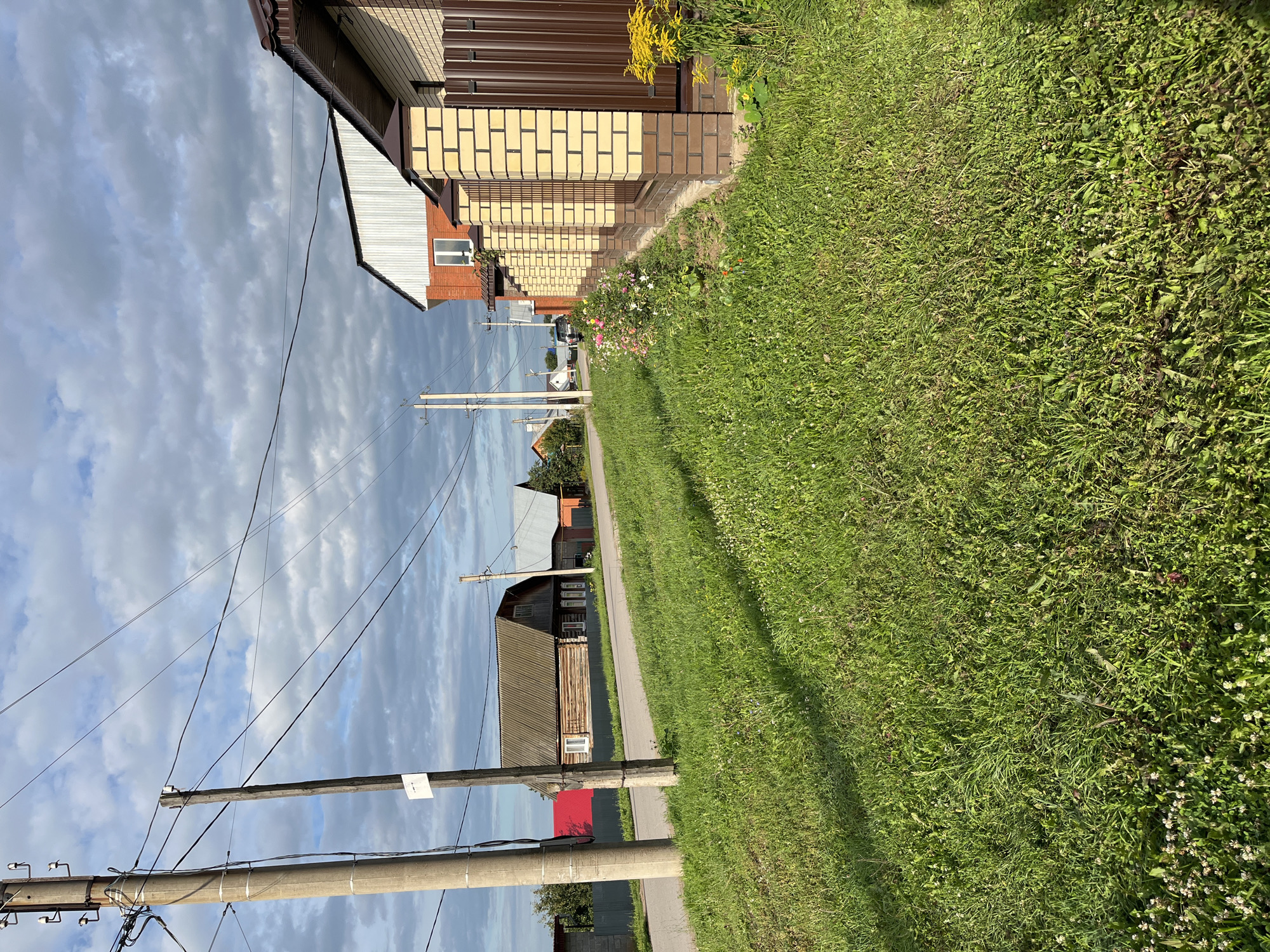 The image depicts a rural or suburban street scene in Ufa, Russia, under a cloudy sky. In the foreground, a well-maintained grassy area lines the side of a paved road. To the right, a fence constructed of light-colored brick with brown metal infill runs along the property line. Adjacent to the fence, a flower bed contains various colorful blooms, including pink and white flowers, and a tall cluster of yellow flowers.
Further down the road, several houses are visible. One house on the right is a modern structure with a red brick lower story and a white, ribbed upper facade. On the left side of the road, older wooden houses with dark roofs are present. One of these houses appears to have a bright red roof. Utility poles with overhead wires are positioned along the roadside, indicating the presence of electricity and possibly telecommunications infrastructure. The environment appears to be a peaceful residential area with a mix of traditional and modern housing. No people are visible in the scene.