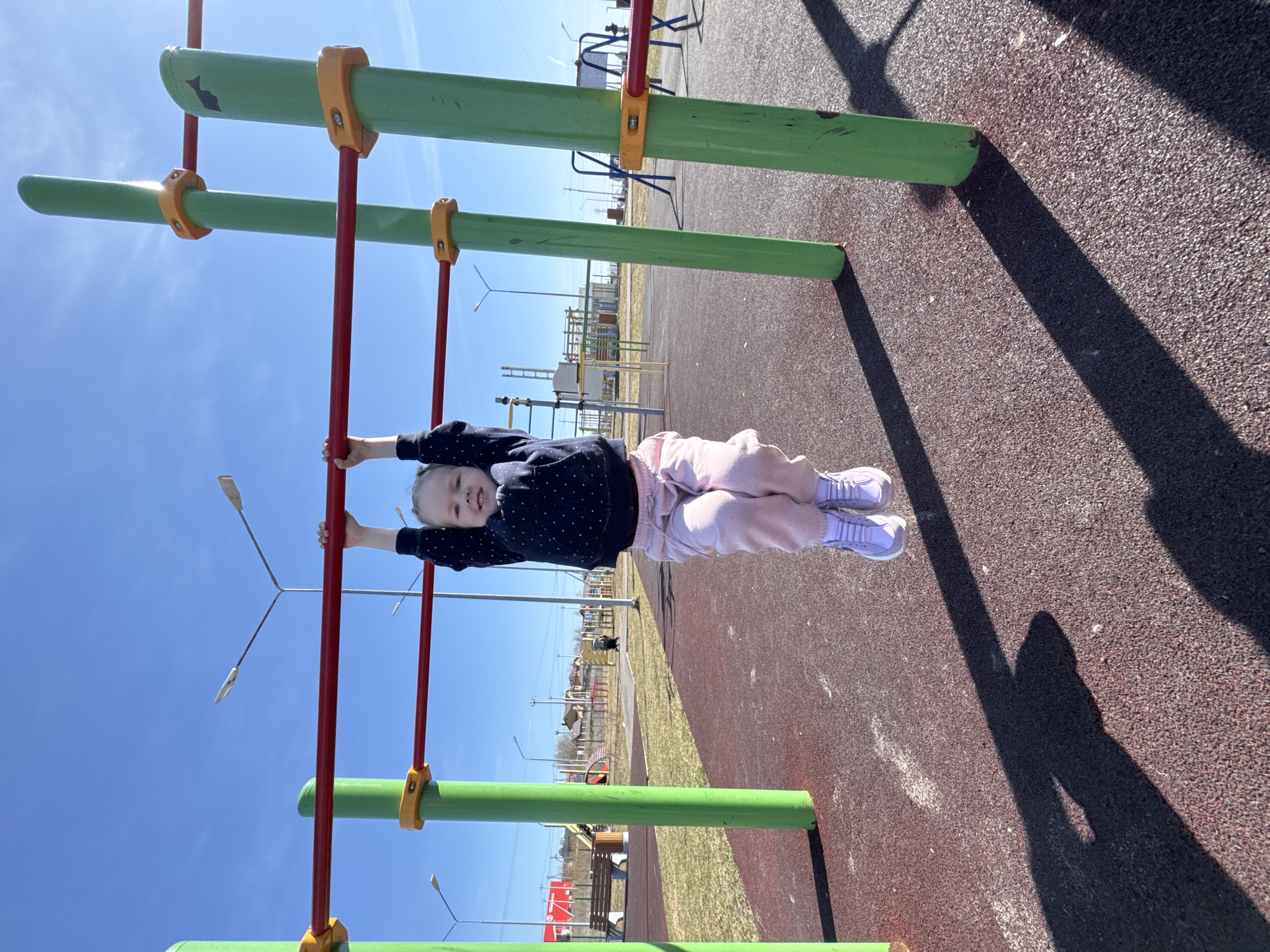A young child is hanging from a green and red monkey bar structure on a playground in Горный, Russia. The playground has a red and brown textured surface. Additional playground equipment and streetlights are visible in the background. The sky is blue. The child is wearing a dark blue shirt with white polka dots, light pink pants, and light purple shoes. The image is taken outdoors in daylight.
