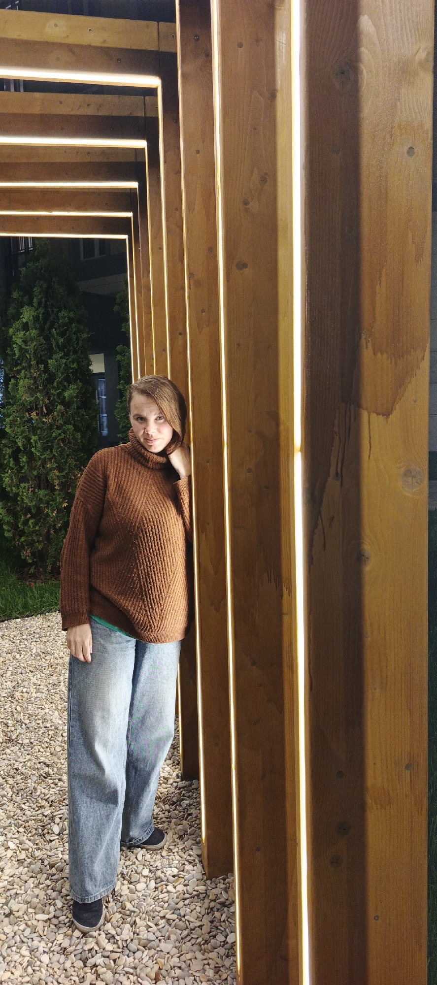 The image displays an adult female standing outdoors at night in the city of Chaikovsky, Russia. She is positioned slightly to the left of center, facing the camera, with her left side leaning against a prominent architectural feature.
The central subject is an adult female with light skin and medium-length, reddish-brown hair parted on the left and swept to her right. She has a neutral to slightly smiling expression and is looking directly forward. Her left hand is raised to her neck area, resting against her turtleneck. She is dressed in a dark brown, chunky knit, high-neck sweater featuring a chevron-like rib pattern on the front. Beneath the sweater, a sliver of a light green garment is visible. Her lower body is clad in light blue, loose-fitting, wide-leg denim jeans, and she wears dark blue or black slip-on shoes.
The architectural feature dominating the right side of the frame and extending into the upper left is a series of light brown wooden beams arranged vertically and horizontally. Each beam incorporates a continuous, thin strip of warm-toned LED lighting along its inner edge, illuminating the structure. The wood shows distinct grain patterns, occasional knots, and visible screw heads. One wider vertical plank on the far right exhibits several dark, vertical stains, suggesting water damage or discoloration. The structure forms a repetitive, receding tunnel-like passage.
The ground beneath the woman's feet and extending into the foreground is covered with light-colored, irregular gravel. To the far left, behind the woman and the wooden structure, a dense, dark green conifer tree is visible. Beyond the tree, in the upper left, parts of a dark building with illuminated rectangular windows are discernible, indicating an urban setting at night. To the far right, beyond the wooden structure, a patch of vibrant green grass is present.