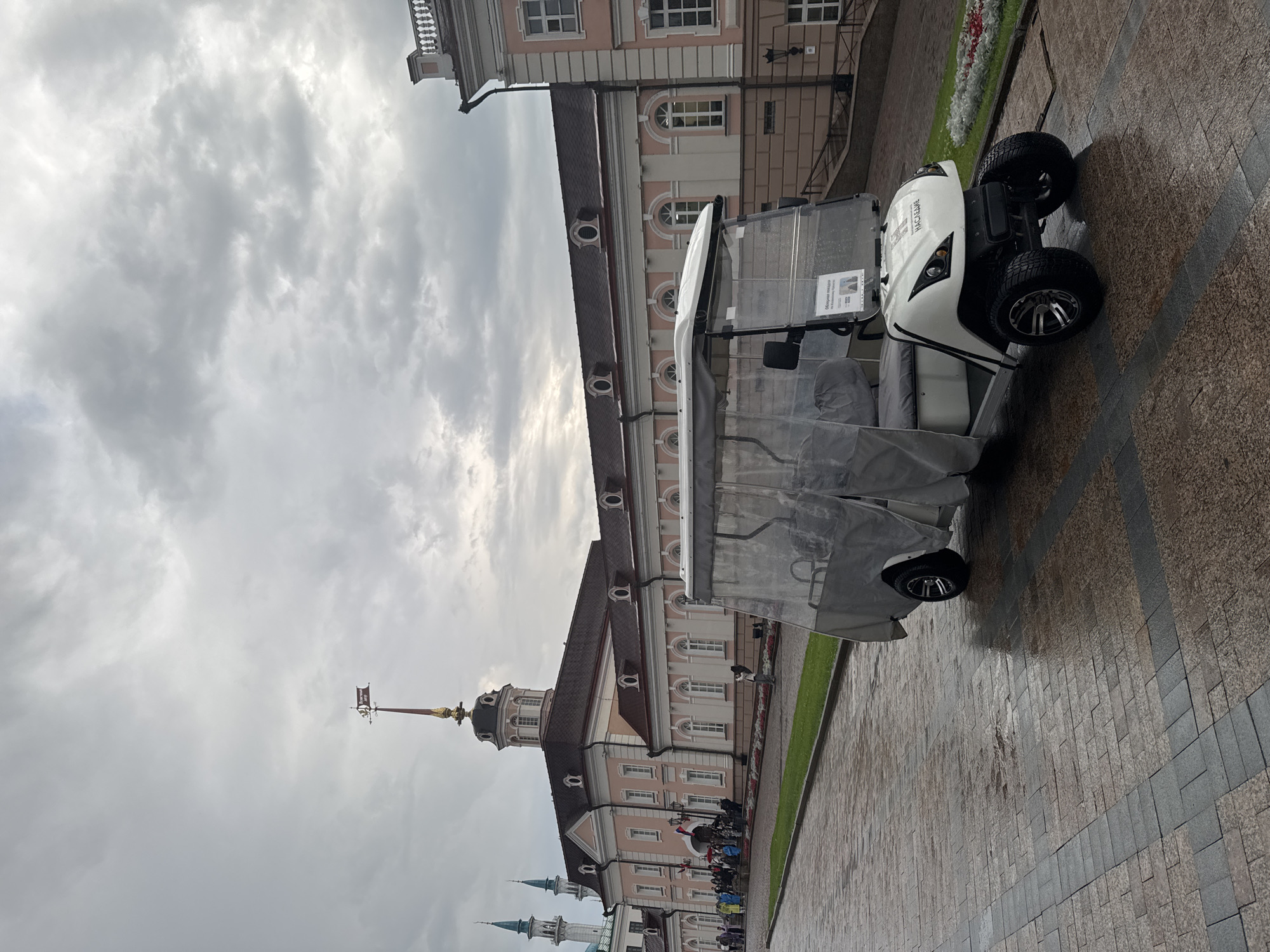 The image depicts a white golf cart with a clear plastic cover parked on a wet, cobblestone-like walkway. The golf cart is positioned in the foreground, angled slightly towards the viewer. Behind the golf cart, a pale pink building with white trim and a dark roof is visible. The building features multiple windows and a central dome topped with a weather vane. To the left of this building, two slender, green-topped towers rise into a cloudy sky. A group of people, some wearing colorful attire, are gathered on the steps leading up to the building. The scene suggests an overcast day, with the wet ground indicating recent rain. This location is the Annunciation Cathedral and the adjacent Kul Sharif Mosque in the Kazan Kremlin, Kazan, Russia.