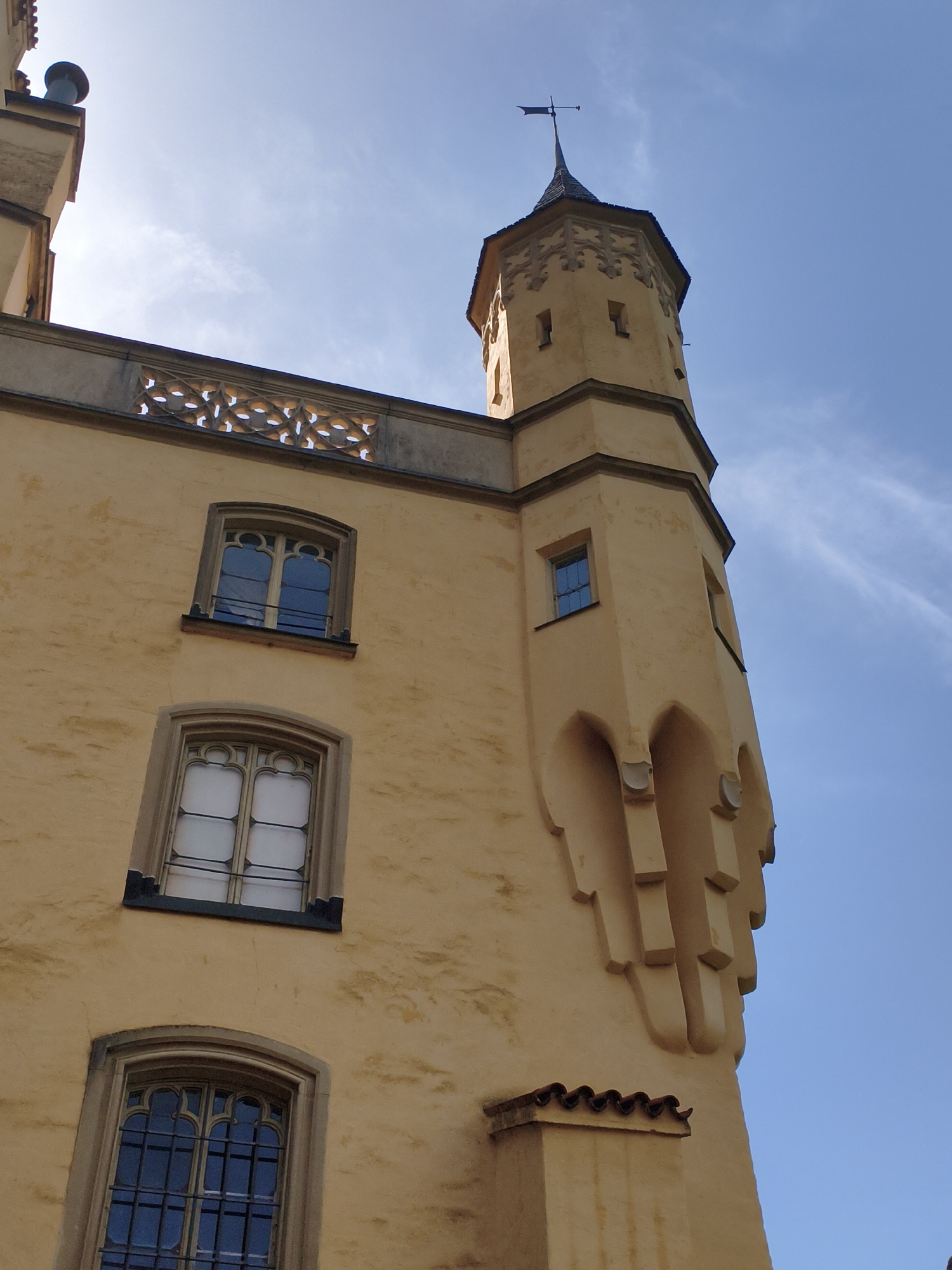 The image shows a close-up, upward-facing view of the exterior of Hohenschwangau Castle in Schwangau, Germany, on a clear blue day. The castle walls are a pale yellow color. On the left side of the image, there are three windows visible on the main building. The topmost window is arched and divided into panes by thin dark mullions. Below it, another identical arched window has its panes covered with a white material. The lowest visible window on this section is also arched and has dark, ornate metal bars over its panes. A decorative stone frieze runs along the top of this section of the castle.
To the right, a tall, octagonal tower dominates the frame. This tower is also pale yellow and features decorative architectural elements, including pointed arches and raised rectangular blocks along its upper section. A dark, pointed spire tops the tower, with a weather vane at its apex. A small, rectangular window is visible on the tower's side. The sky is a vibrant blue with a few thin, wispy clouds. The image captures the grandeur and architectural detail of the castle against the expansive sky.