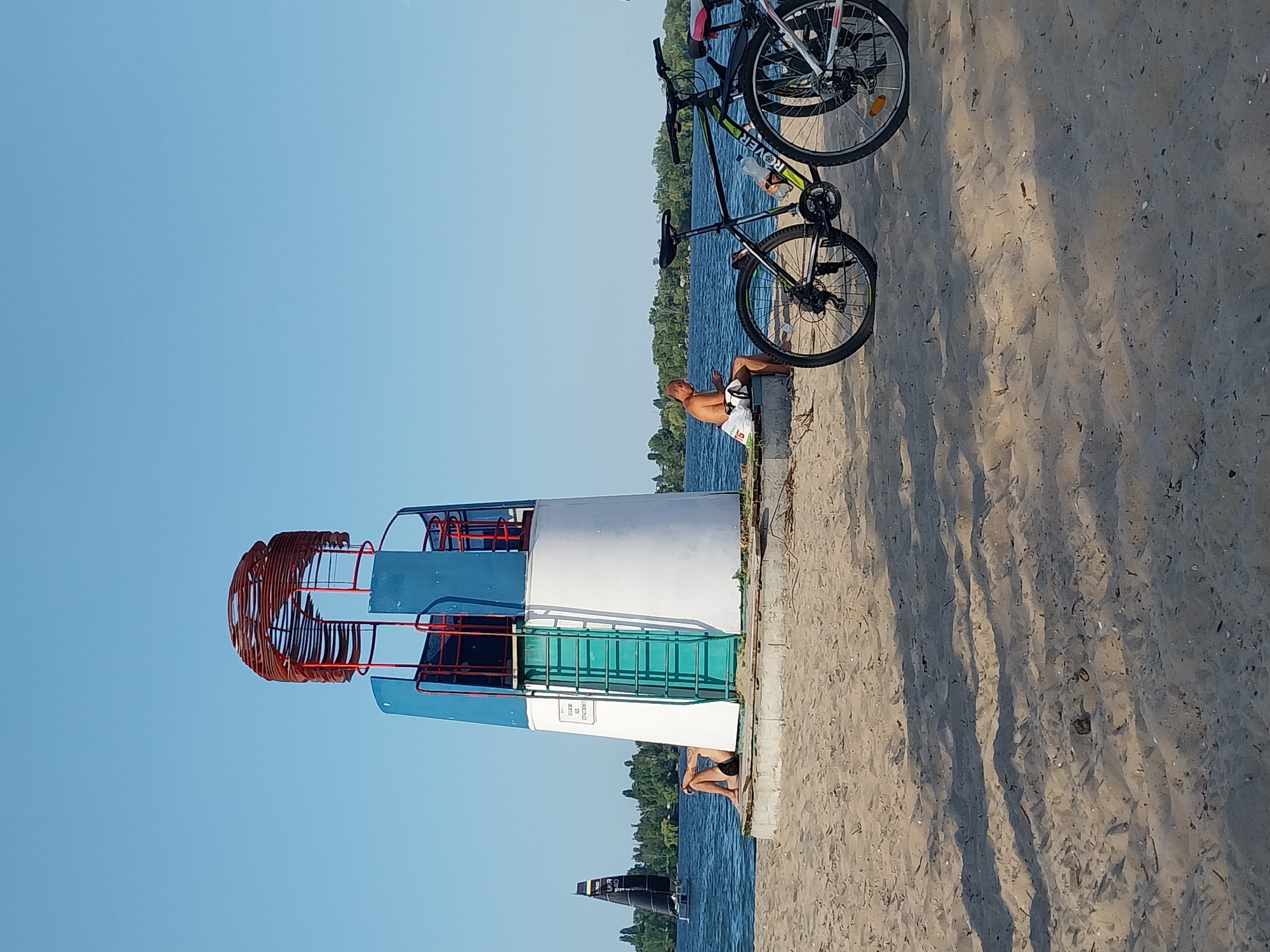 The image depicts a sandy beach along a body of water, likely a river, on a clear, sunny day. In the foreground, two bicycles are parked on the sand. One is black and green with a water bottle attached to the frame, and the other is partially visible behind it, appearing to be white and pink.
Dominating the middle ground is a small lighthouse-like structure. It has a white base, a blue cylindrical section, and a red metal cage-like top. A green ladder is attached to the side of the blue section. A person is seated on a ledge at the base of the lighthouse, facing away from the camera and towards the water, appearing to be looking at a mobile device. Another person is sitting on the sand near the water's edge, to the left of the lighthouse, also facing away from the camera.
In the background, a black sailboat with a white sail is navigating the water. A line of trees and vegetation forms the distant shoreline, suggesting a natural or park-like environment. The scene has a relaxed, recreational atmosphere, common for a summer day at the beach. This location is likely along the Dnipro River in Dnipro, Ukraine, given the presence of a large river and recreational beach areas.