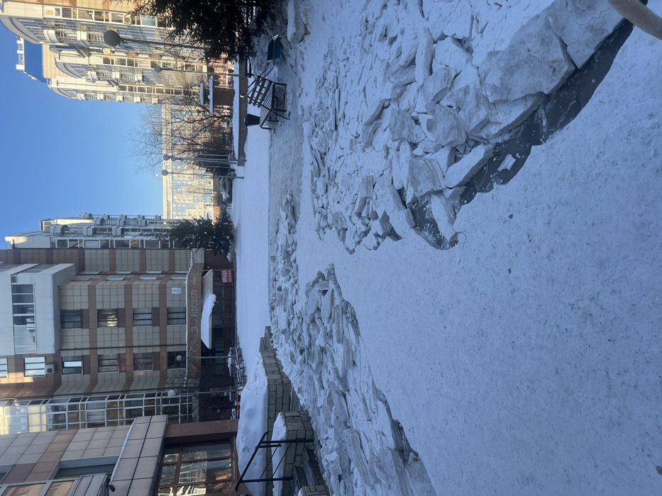 A winter scene shows a sidewalk covered in snow and broken chunks of white material, likely ice or a similar substance. Buildings line either side of the path, with one featuring a storefront. A Christmas tree stands near the building. Benches sit along the path, partially obscured by snow. The sky is clear and blue, suggesting a bright, cold day.