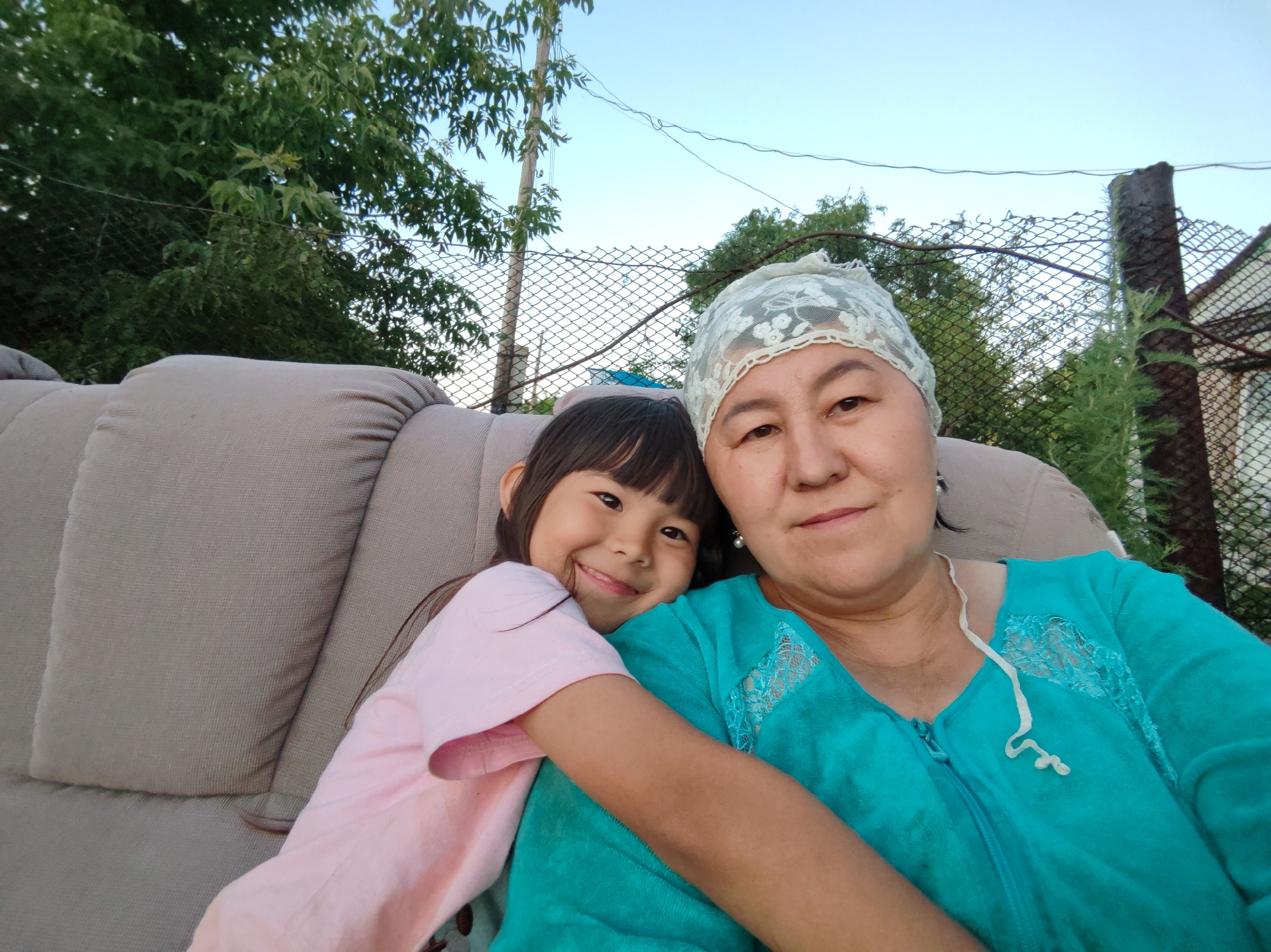 In a candid outdoor selfie, a young girl with dark hair and bangs smiles widely as she hugs a woman. The girl is wearing a pink t-shirt, and the woman is wearing a turquoise top and a white lace headscarf. They are seated on a beige sofa, likely outdoors, with a blurred background of trees, a fence, and a clear blue sky. The overall setting suggests a casual, perhaps residential, environment in the city of Акжар, Kazakhstan.