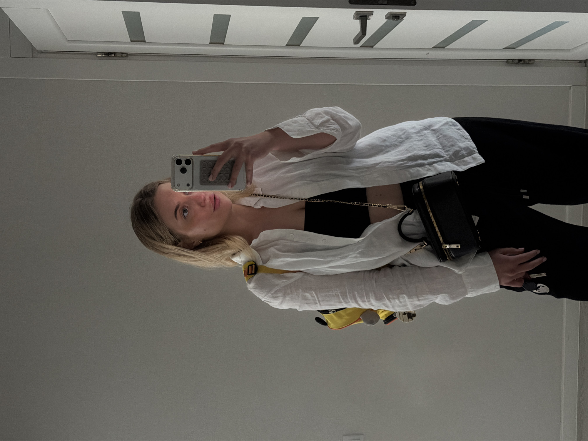 An indoor mirror selfie captures a light-skinned female in what appears to be a residence in Sofiivska Borshchahivka, Ukraine. The subject, with blonde hair, is partially visible from the chest up, looking slightly off-camera to her left. She is wearing an unbuttoned white linen-style collared shirt over a black strapless crop top, with black trousers. A small black rectangular cross-body bag with a gold chain strap is visible on her front. A yellow backpack with grey accents is worn on her left shoulder. She holds a light-colored smartphone with a clear case and a grey textured wallet attachment in her right hand, positioned to take the photograph. Her left hand, down by her side, holds a set of keys, one with a metallic fob. The background consists of a plain light grey wall and a white interior door with two horizontal grey panels, equipped with a silver handle and hinge, visible to the right of the subject.