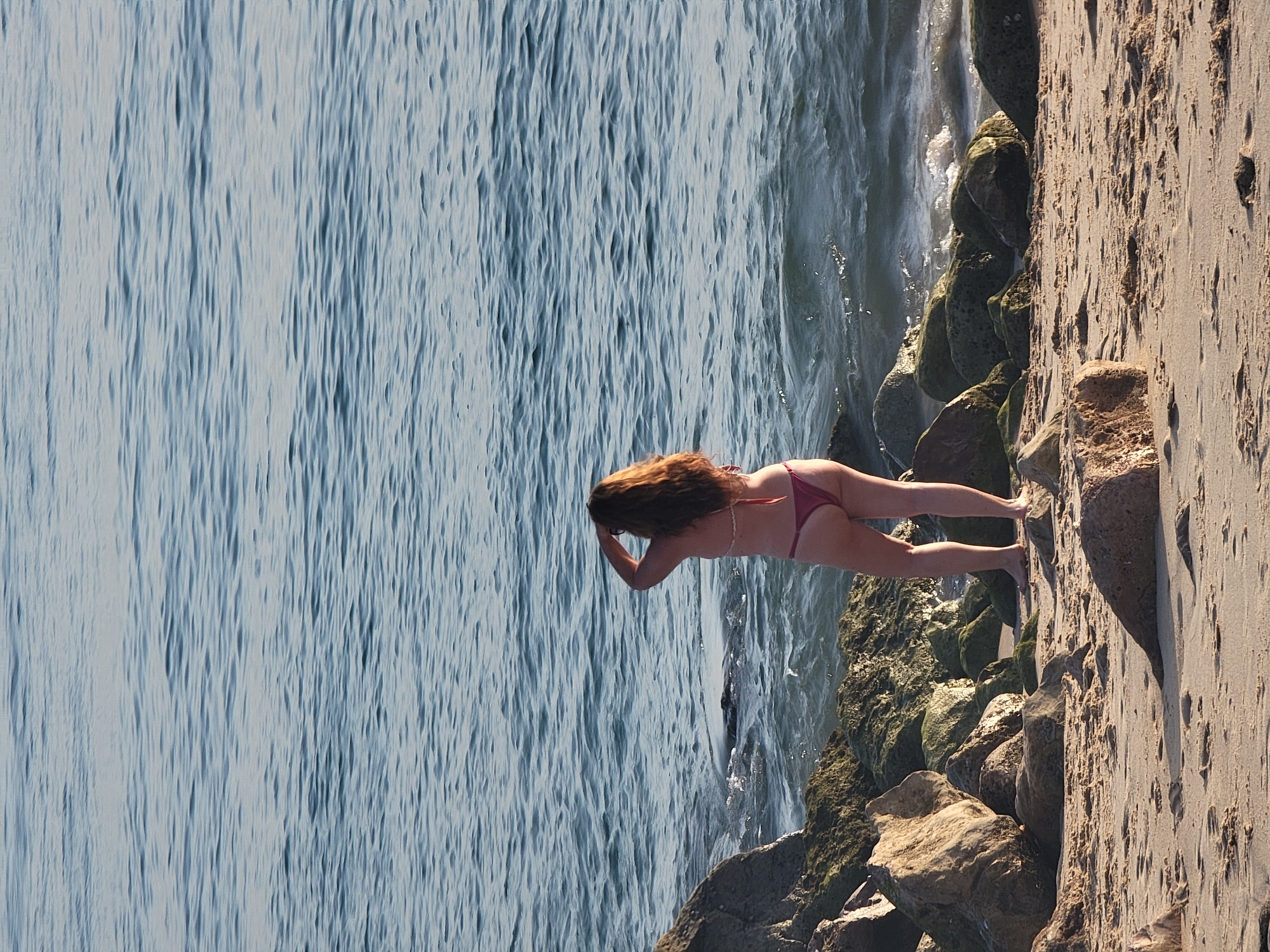 The image shows a woman standing on a sandy beach in Quarteira, Portugal, facing the ocean. She is wearing a dark red bikini and her back is to the camera. Her right hand is raised to her head, obscuring her face. The ocean water is blue and appears somewhat choppy. Rocks line the edge of the beach where it meets the water. The sand in the foreground is littered with small debris and footprints. The lighting suggests it is daytime, likely late afternoon or early morning given the long shadows.