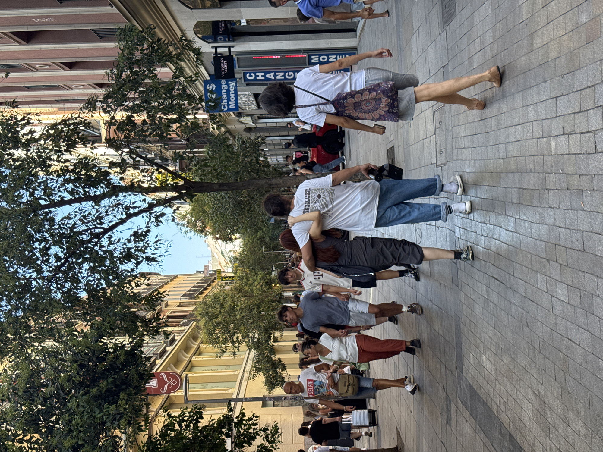The image depicts a pedestrian street scene in Madrid, Spain. In the foreground, a group of people are walking down a paved street lined with buildings and trees. The building on the left appears to be a commercial establishment with yellow awnings and signage, one of which features a red emblem. The building on the right has a more ornate facade with vertical red and beige stripes, and several shops are visible, including one with a sign that reads "Change Money" and "Victoria's Secret".
Several individuals are clearly visible in the midground. A man in a white t-shirt with the word "SELFIE" printed on it, blue shorts, and a crossbody bag is walking towards the camera. Next to him, a woman in a white top and reddish-orange pants is also walking. To their right, two young men are walking away from the camera, one in a grey t-shirt and shorts, and the other in a white t-shirt with a "T" emblem and black shorts. In the background, other pedestrians are visible, as well as a red sports car.
The street is paved with rectangular stone tiles. Trees with full green foliage are planted along the street, providing shade. The sky is visible in the upper part of the image, appearing clear and blue. The lighting suggests it is daytime, with sunlight casting shadows on the street. The overall atmosphere is that of a bustling urban environment with people enjoying a walk.