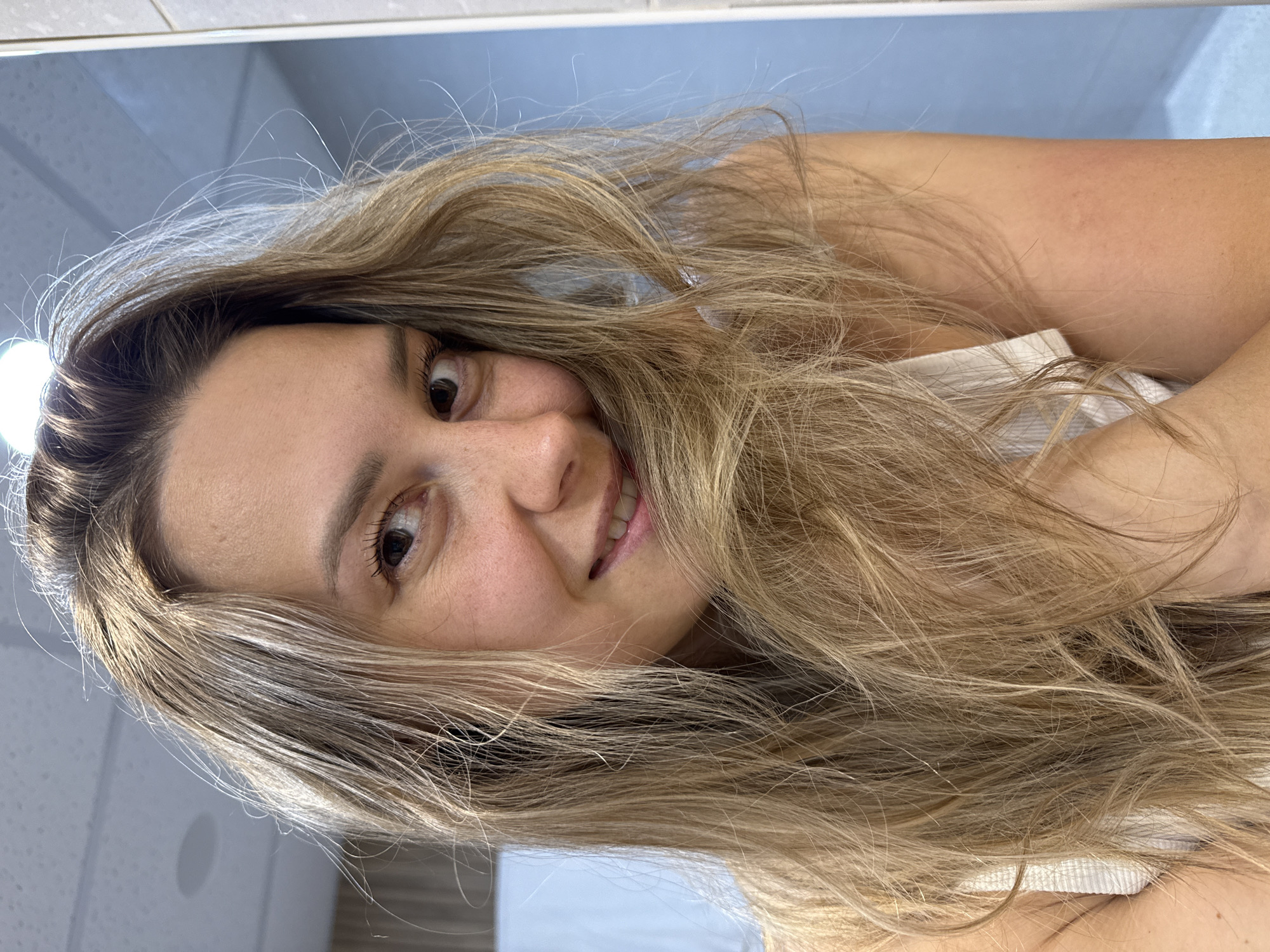 A young woman with long, blonde, wavy hair is captured in a close-up, self-portrait style photo. She is looking directly at the camera with a slight smile, her head tilted. Her hair is framing her face, obscuring part of her left cheek. She appears to be indoors, possibly in a bathroom, with a tiled wall visible on the right side of the image and a ceiling light fixture in the upper left corner. The overall lighting suggests a natural light source or a well-lit interior. There is no indication of the city of Nebug, Russia in the image.