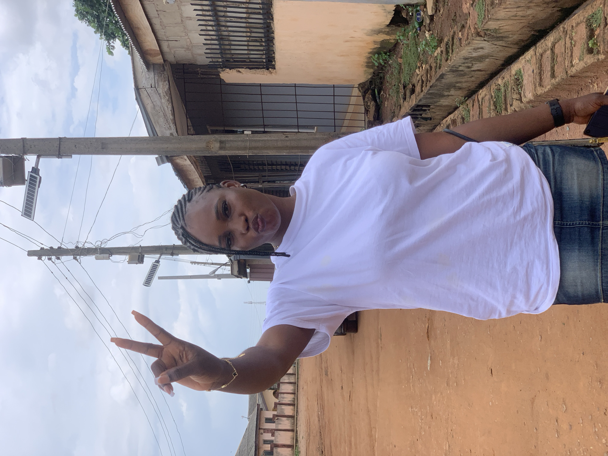 The photograph depicts a woman in Ikorodu, Nigeria, making a peace sign with her left hand. She is wearing a white t-shirt and what appears to be a denim skirt. The background includes a building with barred windows, a utility pole with solar-powered lighting, and a narrow sidewalk bordered by foliage. The overall setting suggests a residential area.