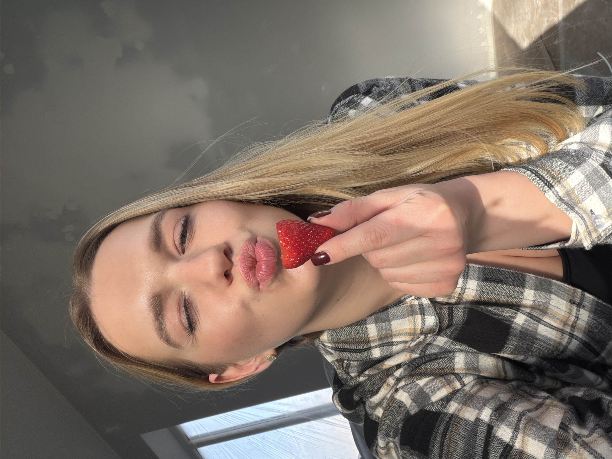 A blonde woman is holding a strawberry to her lips while wearing a black and white checkered shirt. The background shows a cloudy sky, part of a window, and a wall. There is nothing in the image to confirm the location being in Dreiliņi, Latvia.