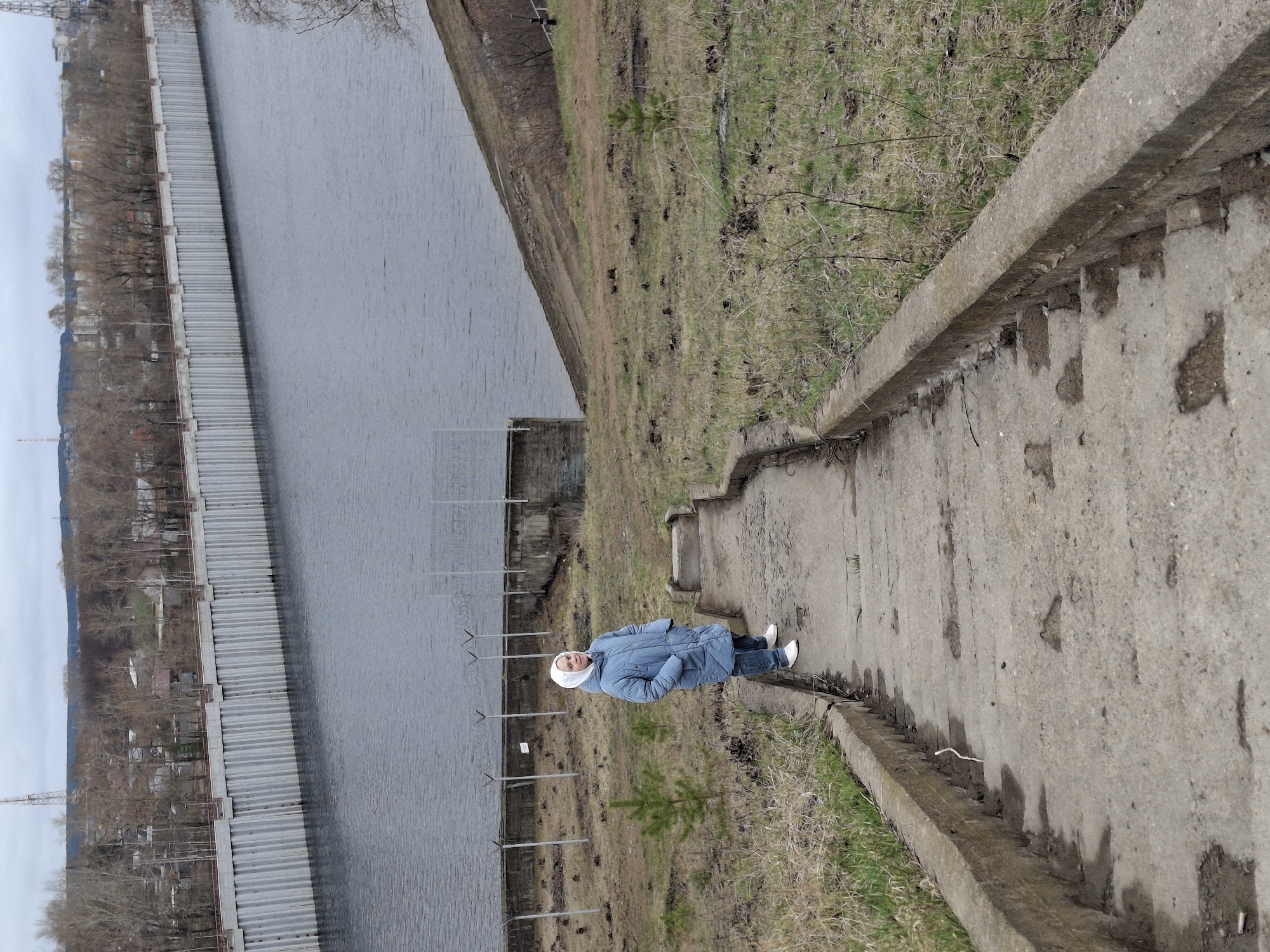 A medium, eye-level shot presents a woman standing on a concrete pathway beside a large body of water. The pathway, with worn, cracked surfaces and bordered by concrete edges, appears to be part of a structure leading down to the water's edge. Patches of vegetation and dry grass line the sides of the pathway.
The woman is seen wearing a light blue jacket, jeans, and white shoes. Her head is covered with a white headscarf. She is positioned in the middle of the pathway, facing the camera.
To the left of the pathway, a long body of water is visible, bordered by a metal barrier along the embankment. Beyond the barrier, a cityscape with buildings and bare trees stretches into the distance. The sky is overcast, suggesting a cloudy day.
This scene is located in the city of Chaikovsky, Russia, as indicated in the instructions. The image captures a typical urban landscape where concrete infrastructure meets natural water bodies and urban development.