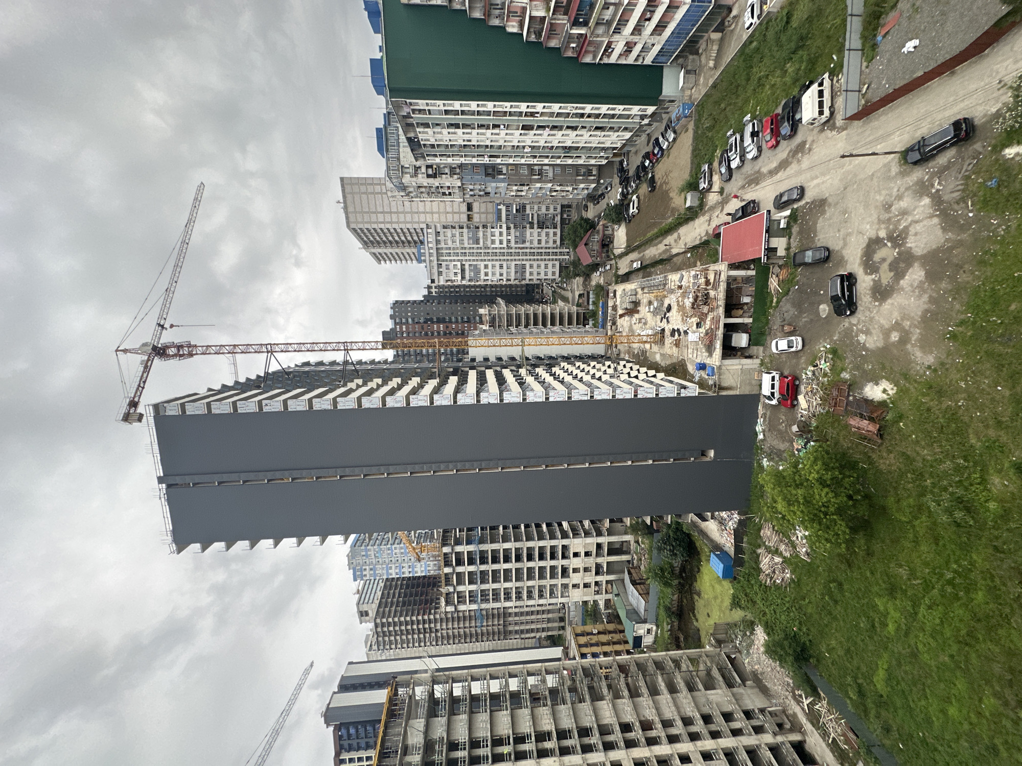 The image depicts an aerial view of a construction site in Batumi, Georgia, characterized by numerous unfinished high-rise buildings and active construction cranes. The scene is dominated by the stark grey facade of a partially completed skyscraper in the foreground, towering over adjacent skeletal structures. Several other buildings in various stages of construction are visible, some with exposed concrete frameworks and others with partially installed facades. A large construction crane with a red and white boom is prominently positioned, suggesting ongoing work.
The ground level reveals a mix of unpaved dirt paths and grassy areas, interspersed with piles of construction debris and lumber. A row of parked cars, including sedans and an SUV, lines a dirt road adjacent to one of the buildings. A small, red-roofed structure, possibly a temporary site office or a small house, is situated amidst the construction zone. The overall environment is overcast, with a cloudy sky contributing to a muted color palette. The image captures the dynamic and ongoing development typical of urban construction projects.